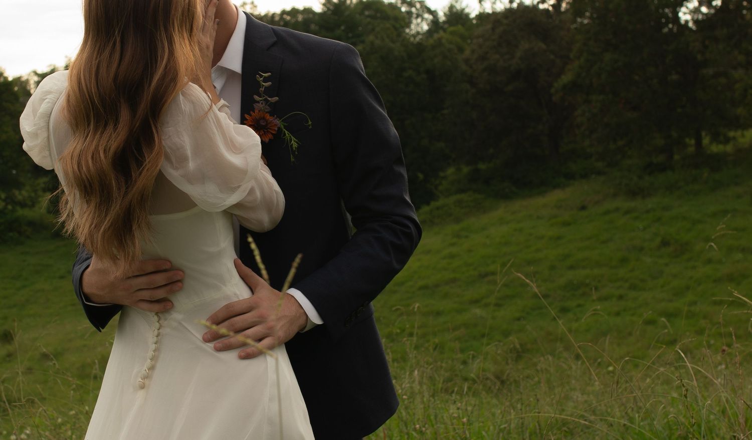 A bride and groom embrace in a lush green field, the bride in a white dress with pearl details.