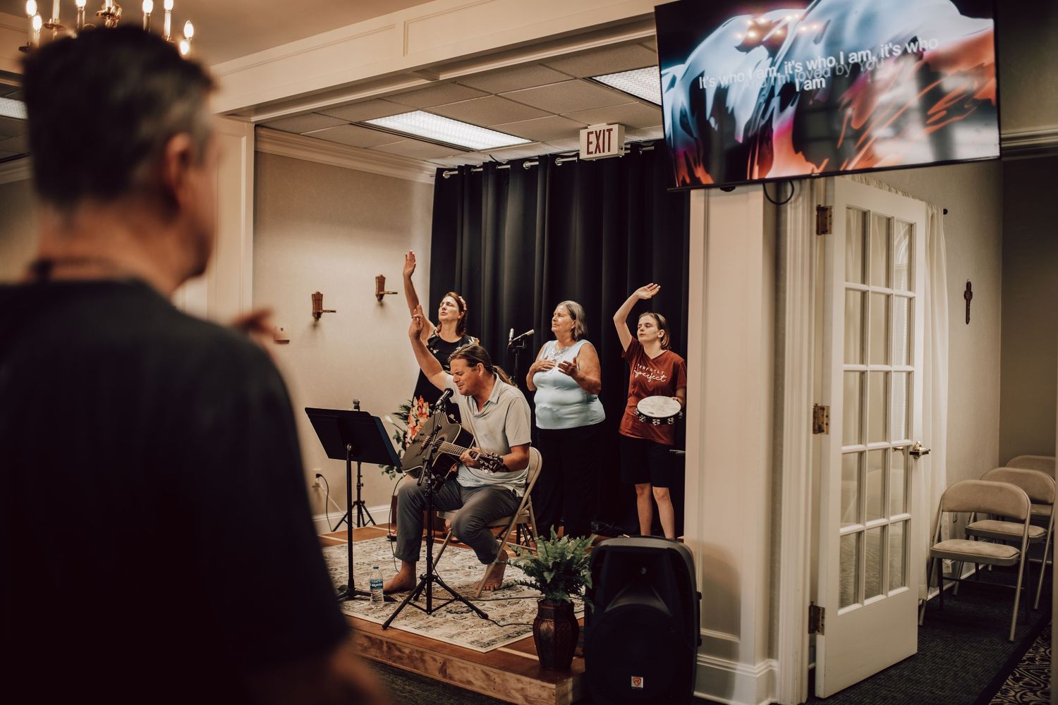 Worship team singing and praising with hands raised during an indoor church service with screen display.