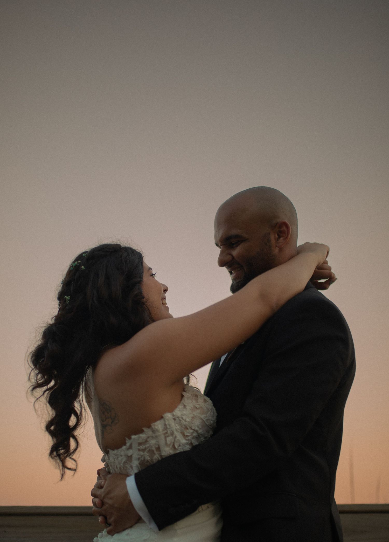 A bride and groom share a romantic embrace at sunset, smiling at each other in wedding attire.