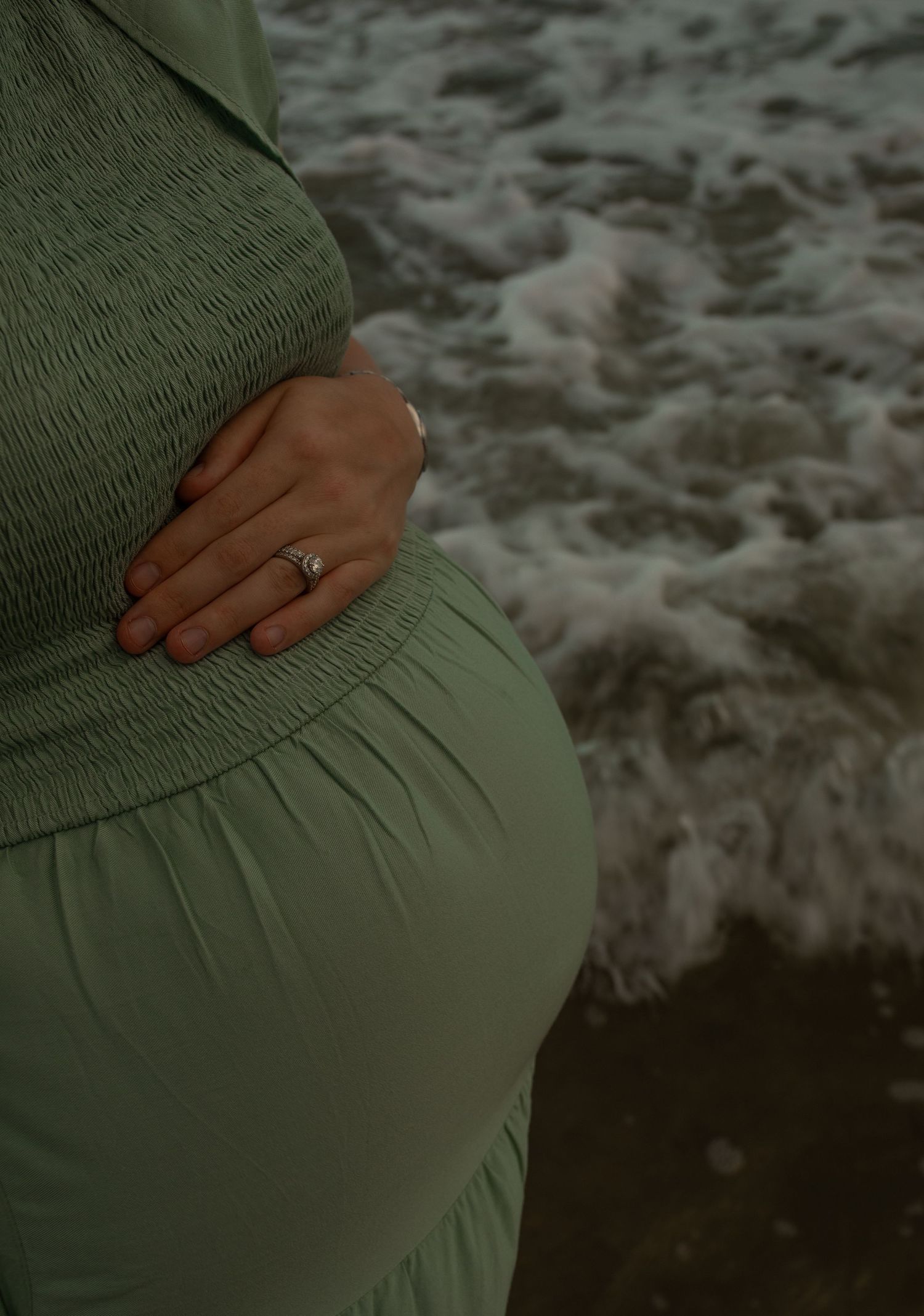Hand resting on a pregnant belly in a sage green maternity dress against a blurred ocean background.