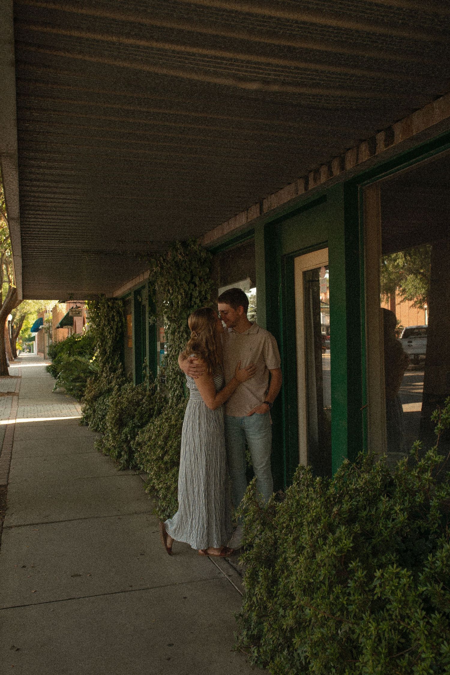 A couple stands close together under the covered walkway of a green-trimmed building with bushes at golden hour.