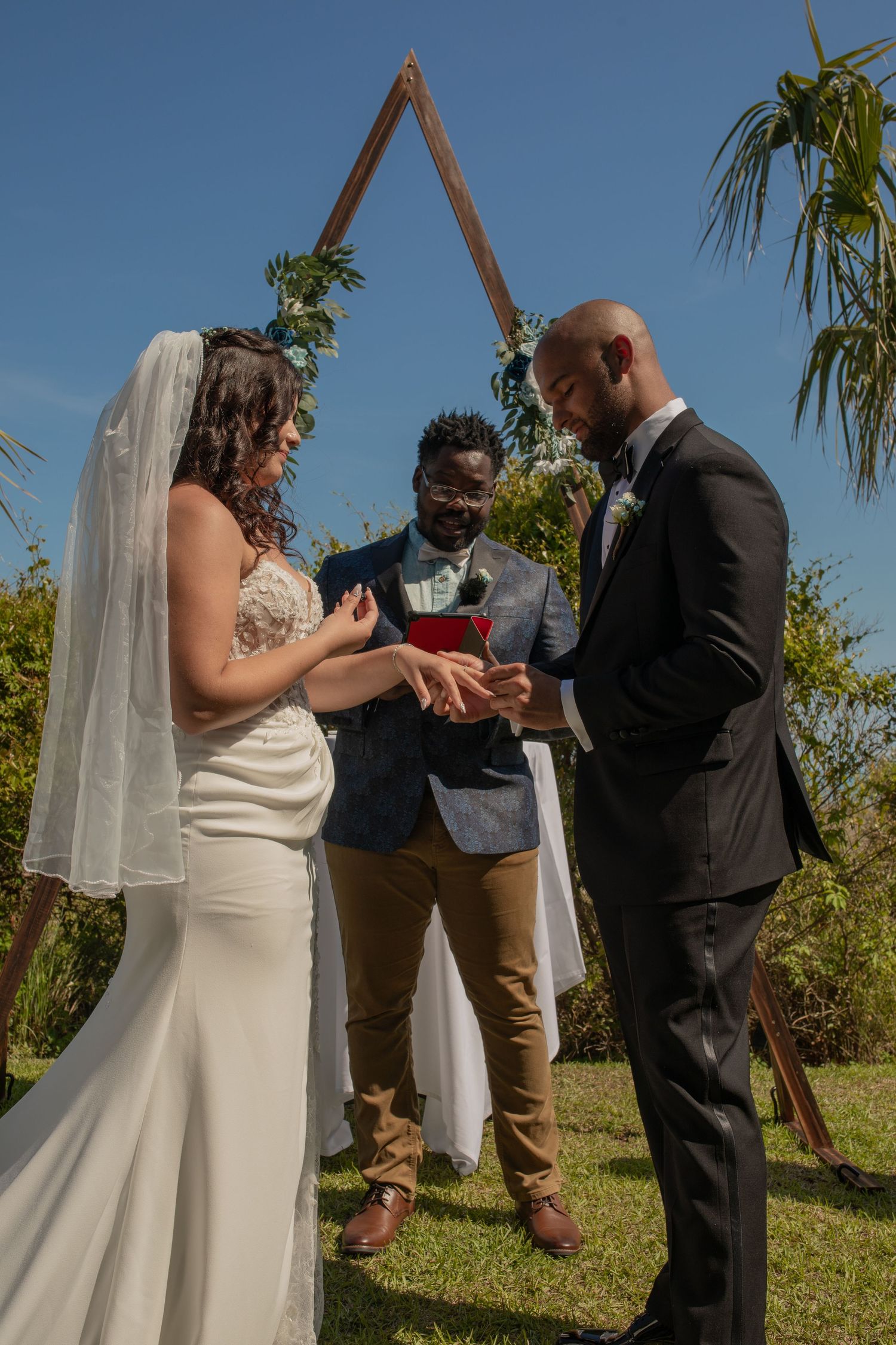 Interracial couple exchanging wedding rings outdoors under triangular arch with officiant, blue sky background.
