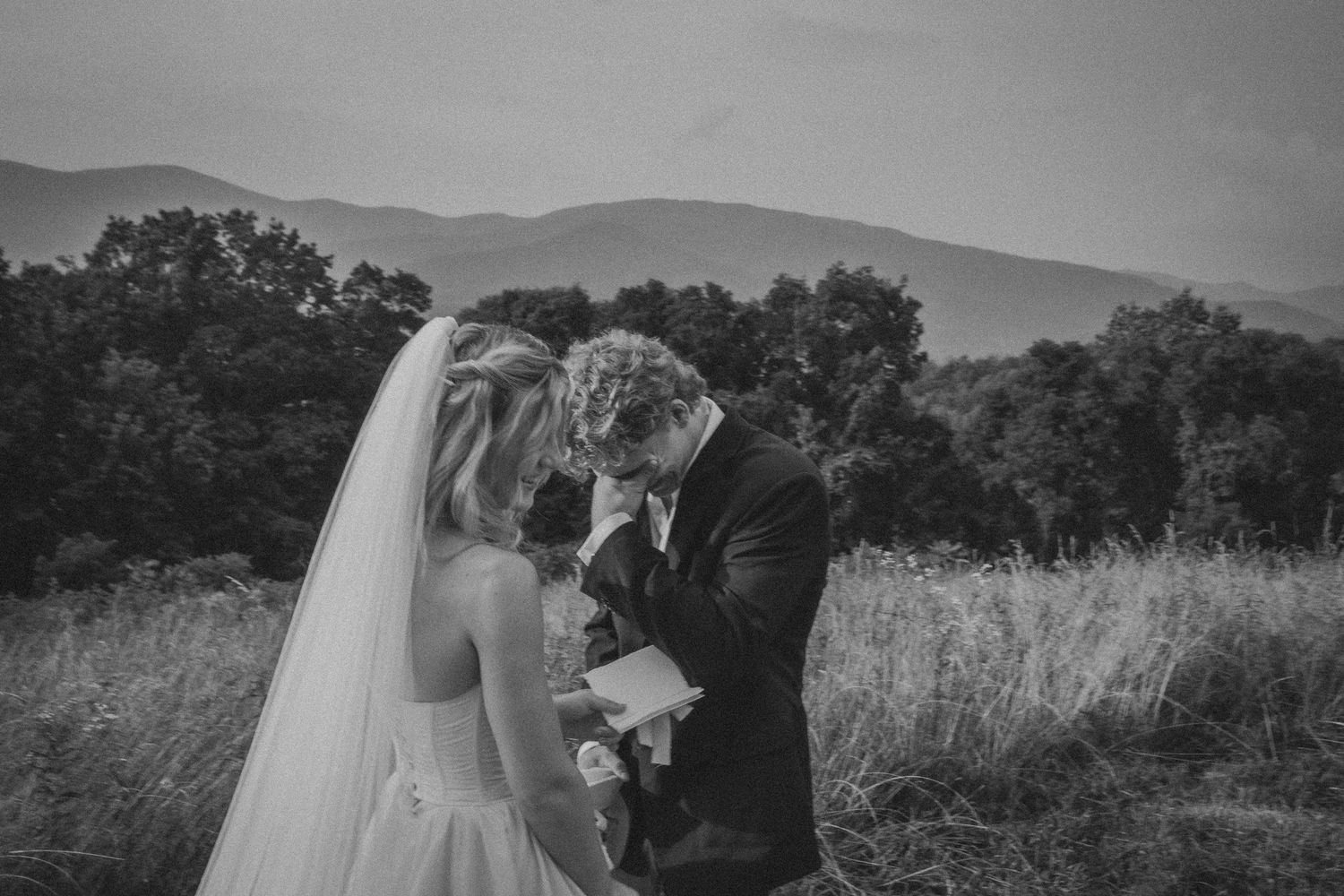 Black and white photo of a bride and groom sharing an intimate moment in a field with mountains in the background.