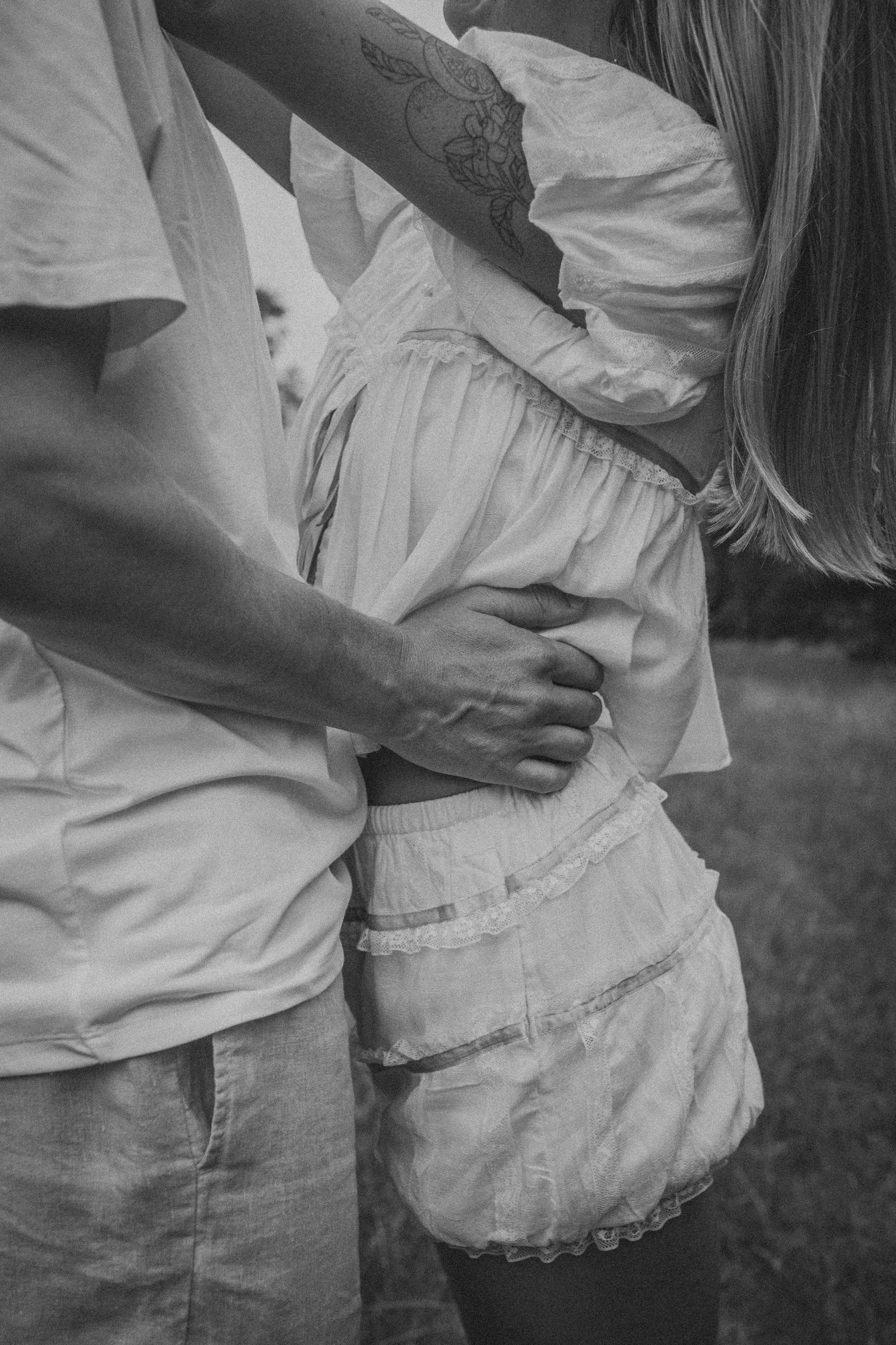 Black and white photo of a couple embracing, man's hand on woman's waist, woman wearing white dress with floral tattoo.