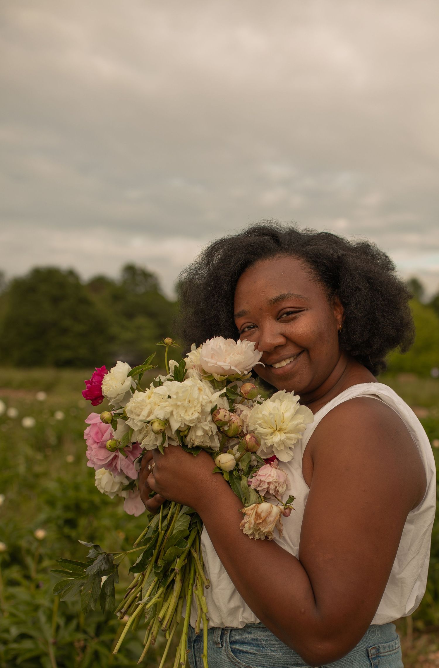 Smiling woman holding a bouquet of white and pink peonies in a flower field on a cloudy day.