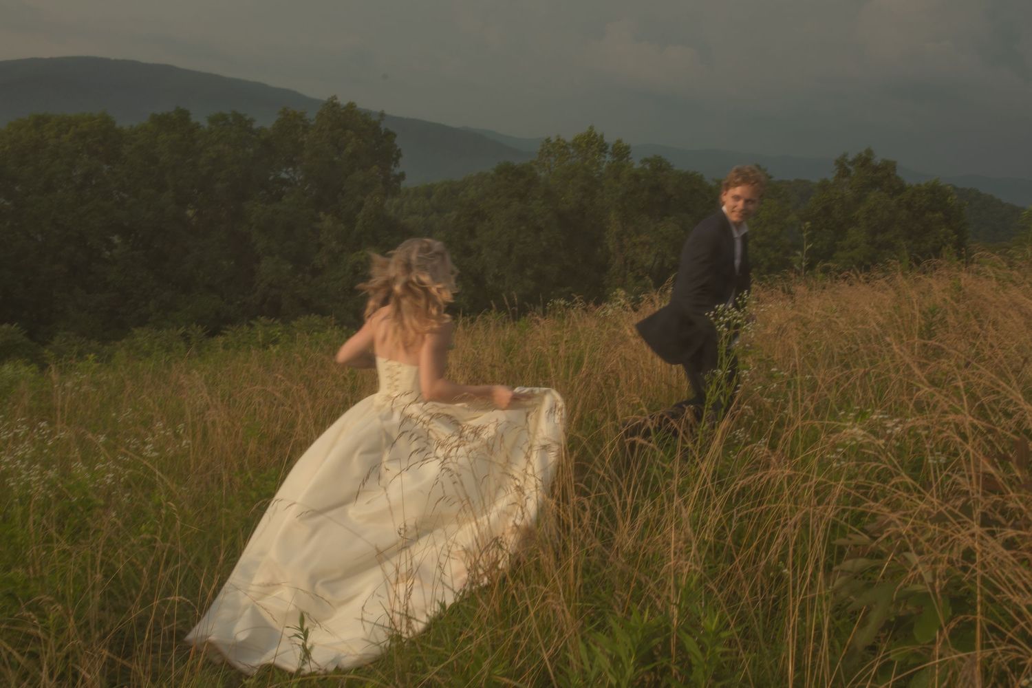 Bride in white gown and groom in suit walk through tall grass field with mountain backdrop on wedding day.