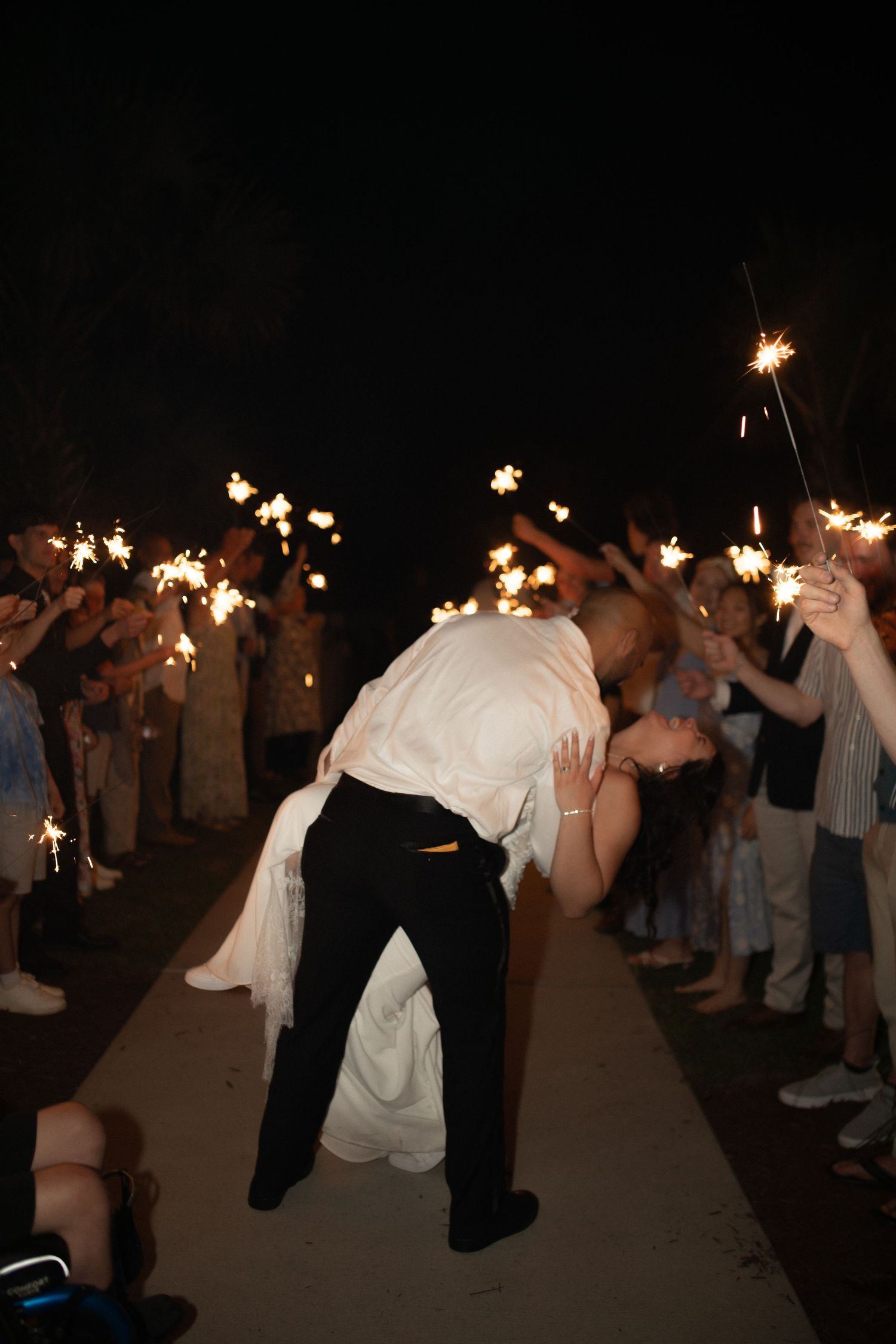 Bride and groom share romantic dip kiss during sparkler send-off exit at night wedding celebration.