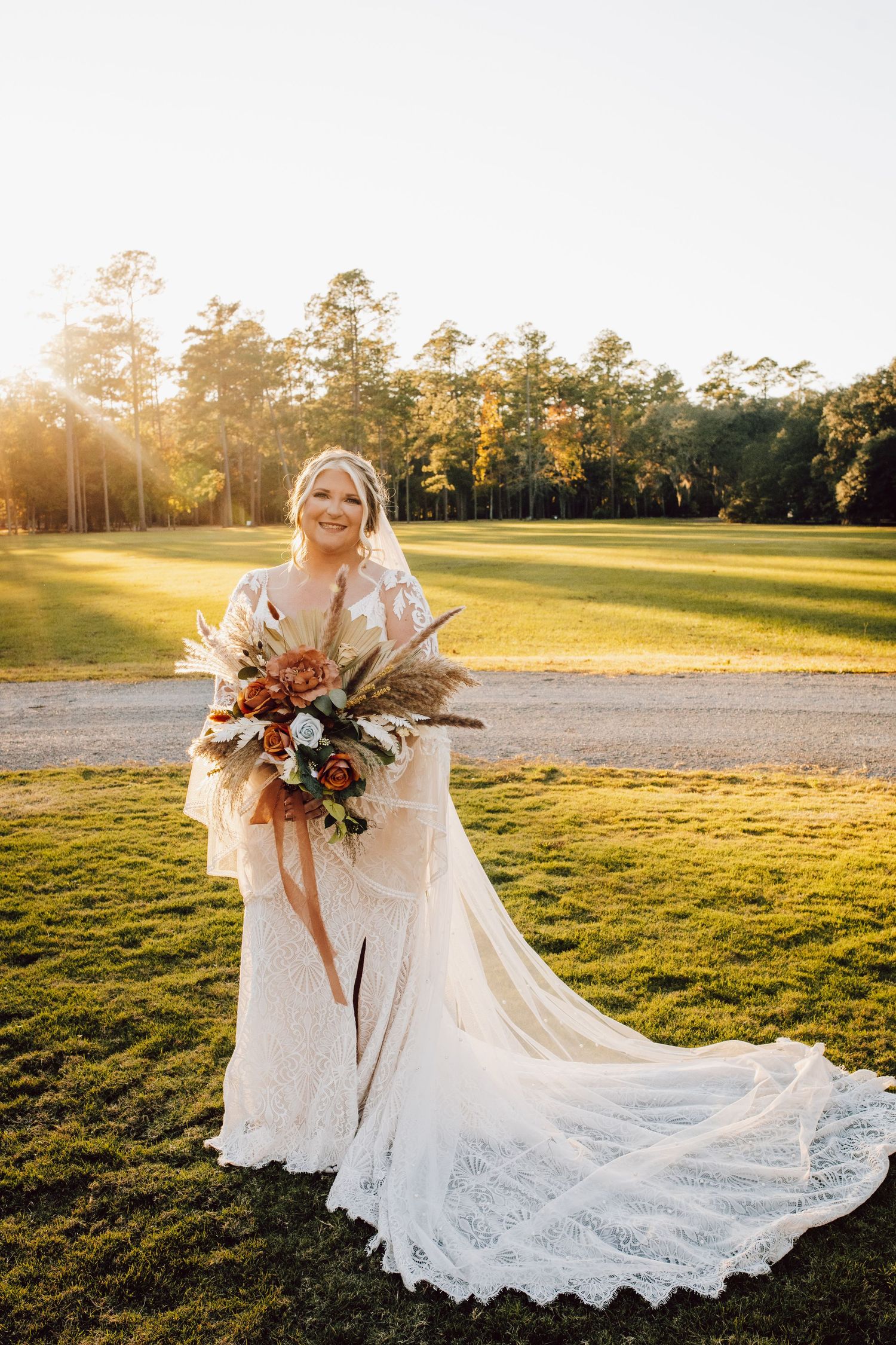 A bride in a flowing lace wedding dress holds a rustic bouquet on a sunlit golf course at golden hour.