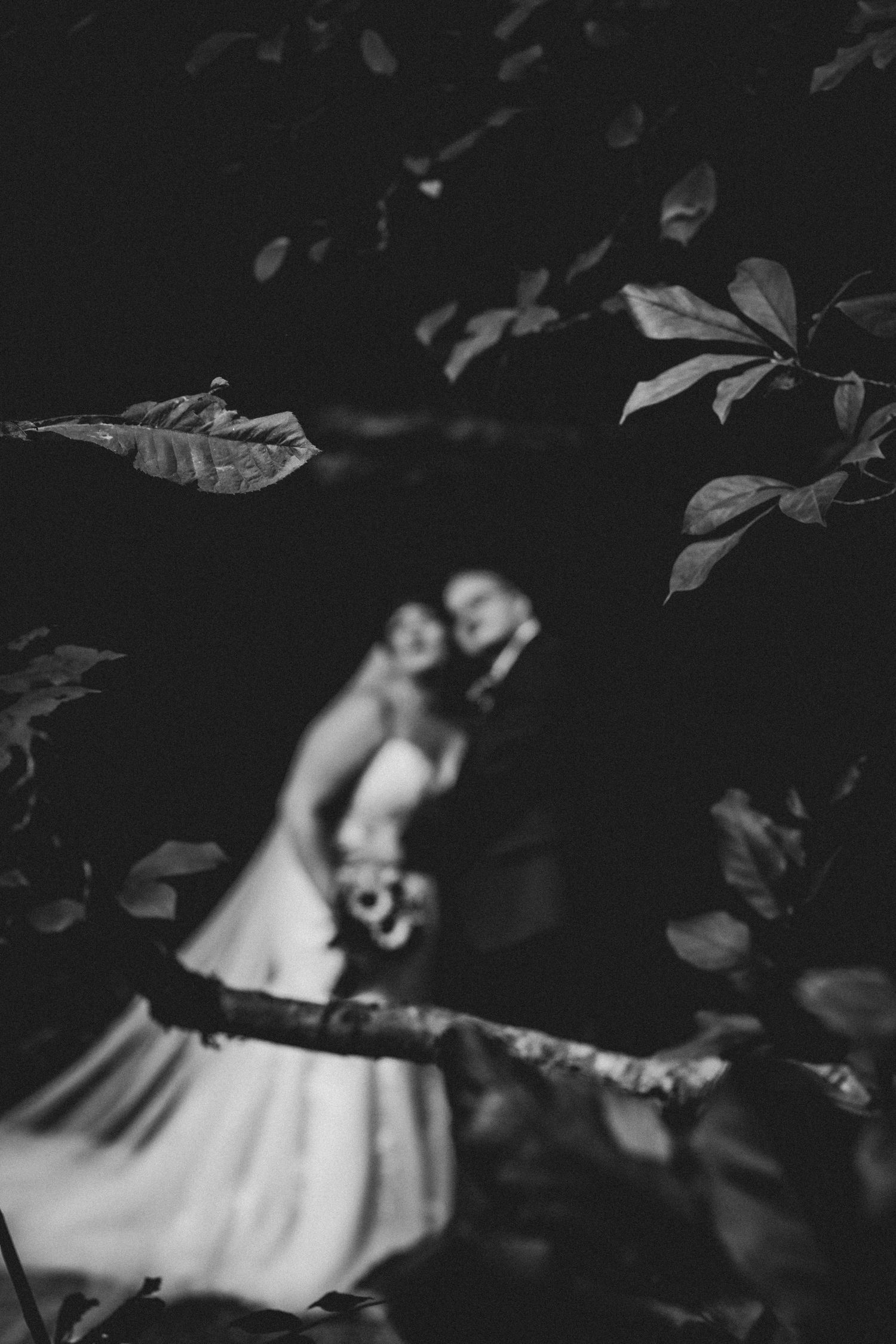 Black and white wedding photo of bride and groom framed by blurred leaves and branches in a romantic garden setting.