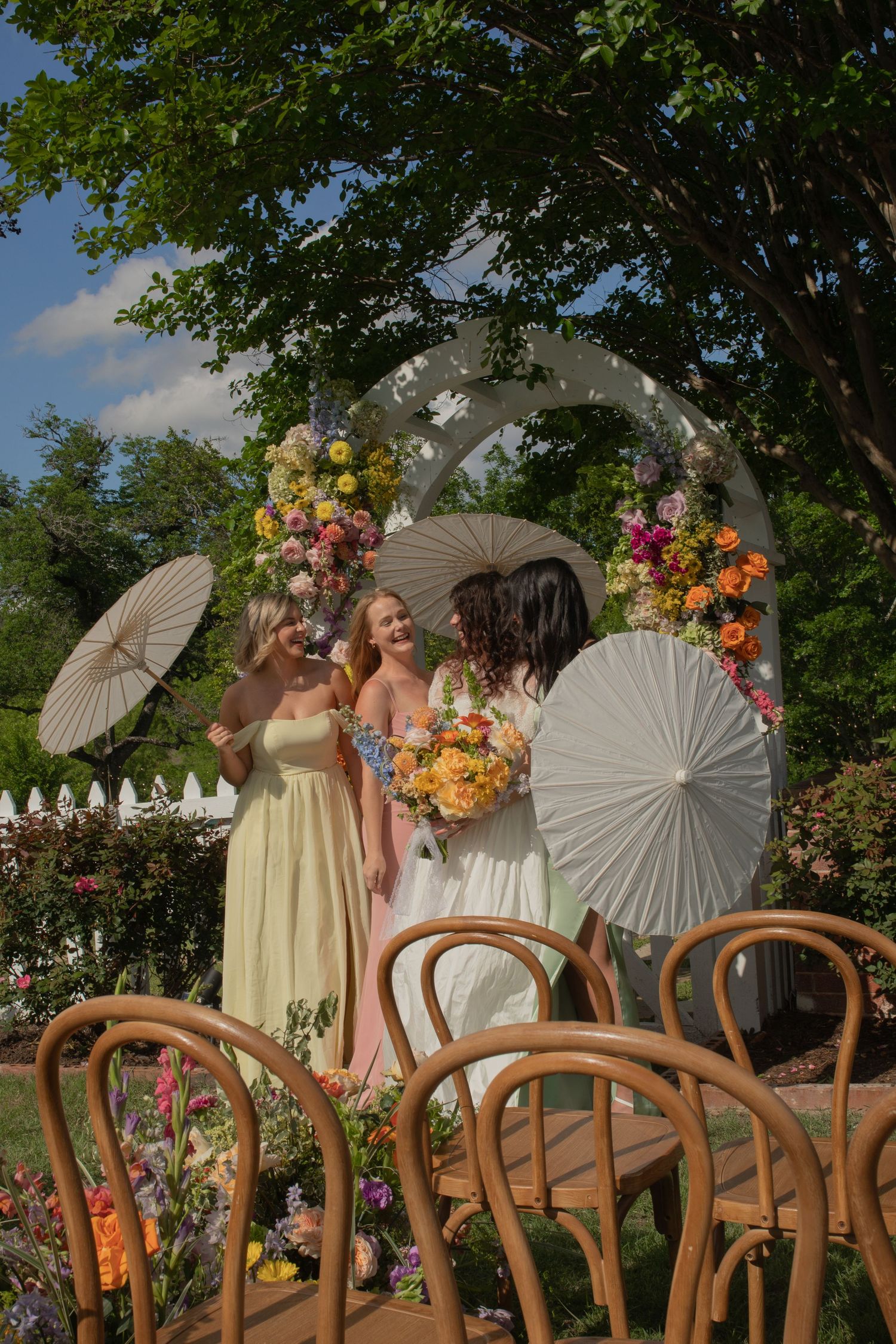Bridesmaids in pastel gowns holding parasols stand by a floral arch in a garden wedding setting.