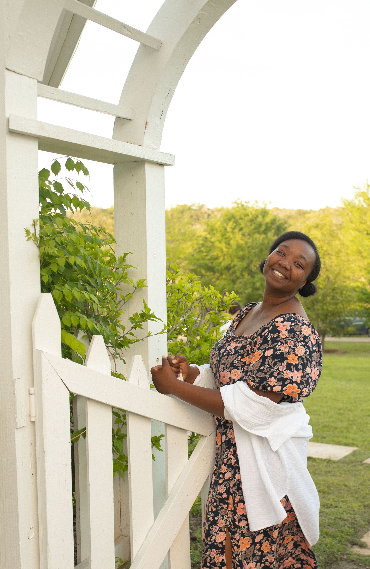 Smiling woman in floral dress holding white jacket by a white garden arbor gate with green plants and trees.