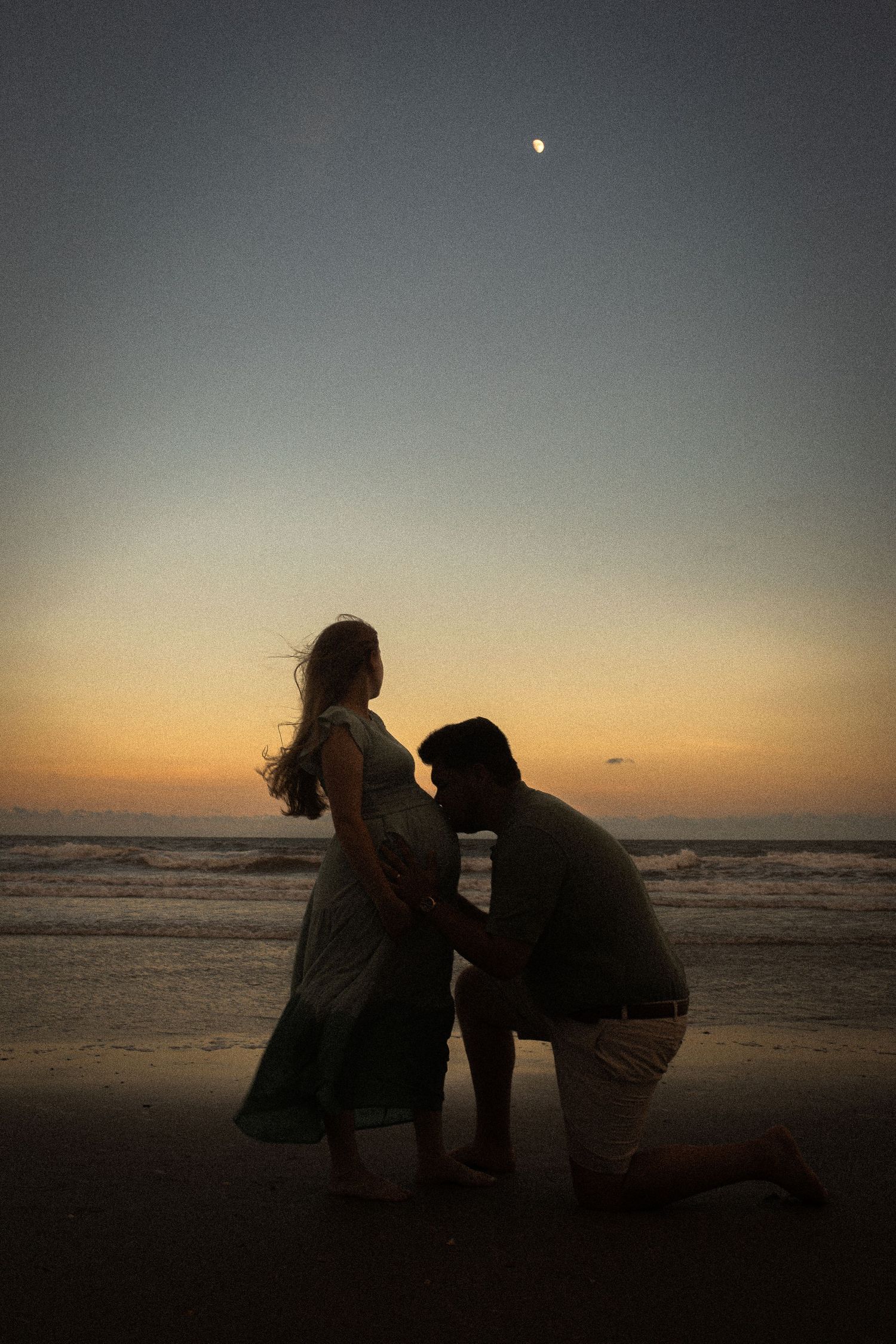 A silhouetted couple shares an intimate moment on the beach at sunset with the moon visible in the dusky sky.