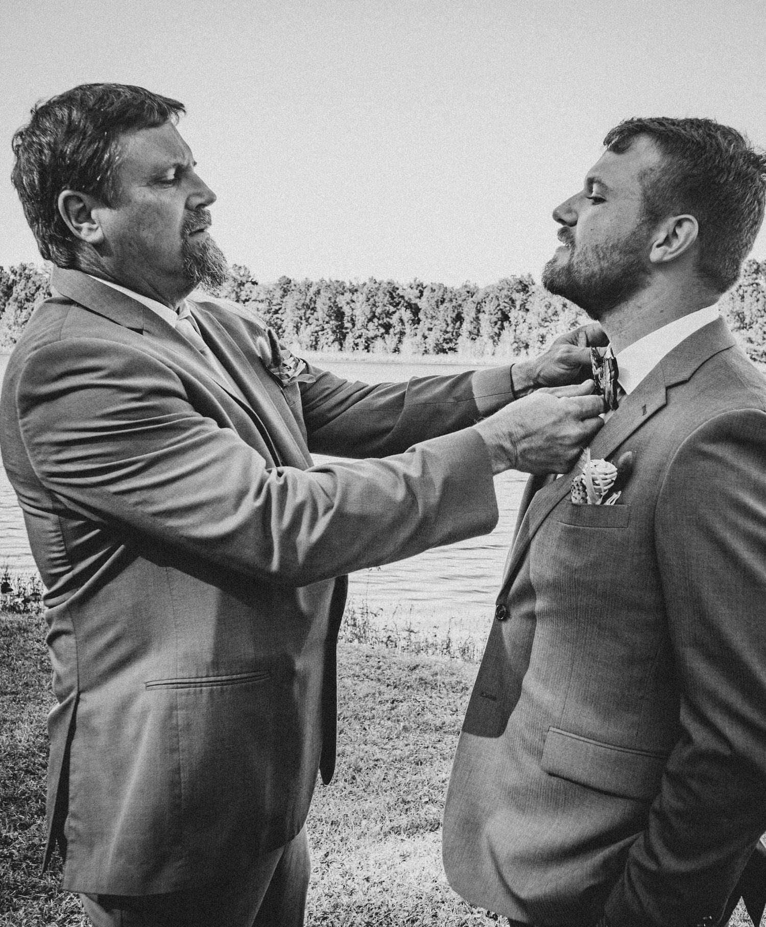 Two groomsmen in gray suits adjust each other's boutonnieres during an outdoor wedding photoshoot in black and white.