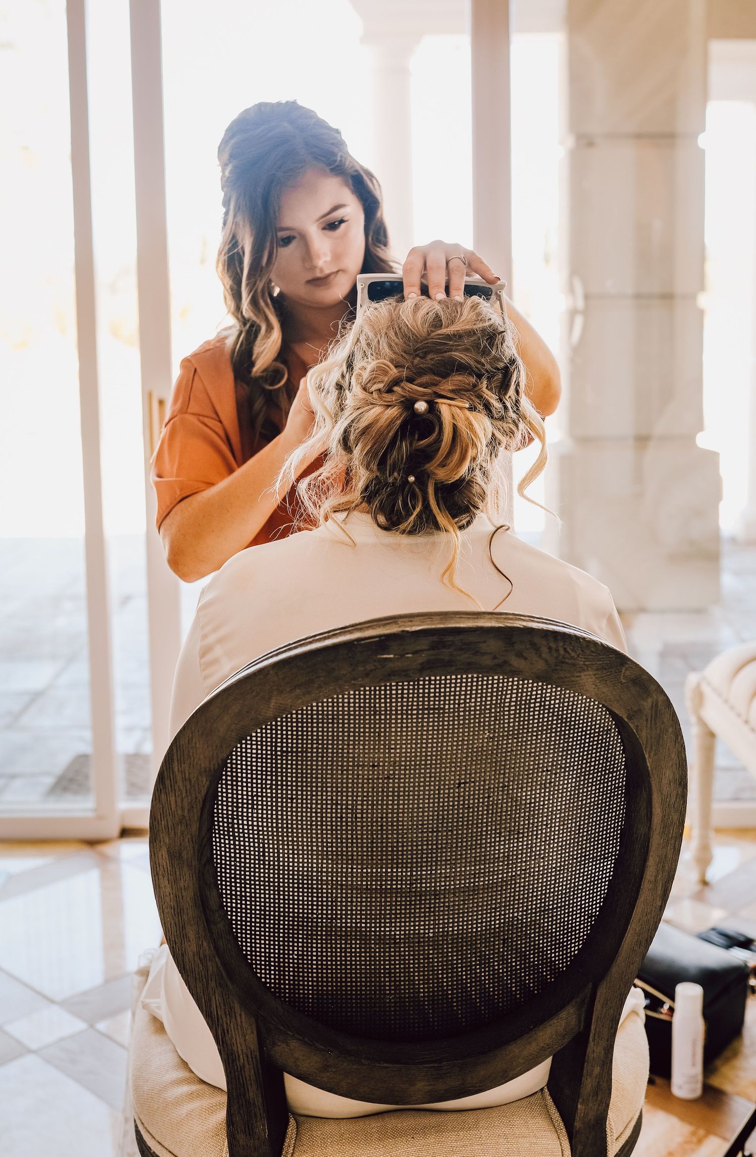 A hairstylist styles bride's elegant updo with cascading curls in a sunlit room with vintage chair.