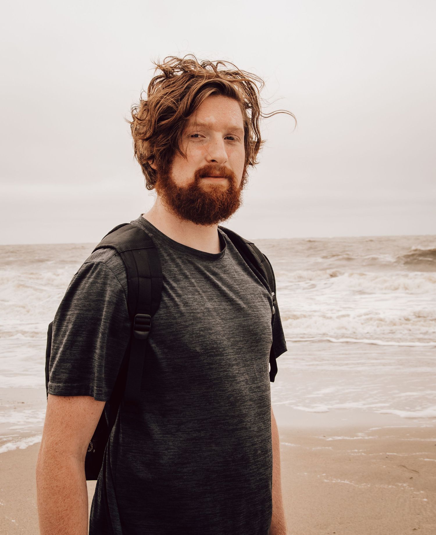 Person wearing dark shirt with tousled hair and beard standing on beach.