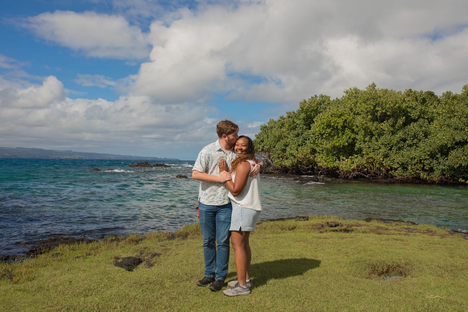 A couple shares a romantic moment on a lush green shoreline overlooking turquoise waters in Hawaii.