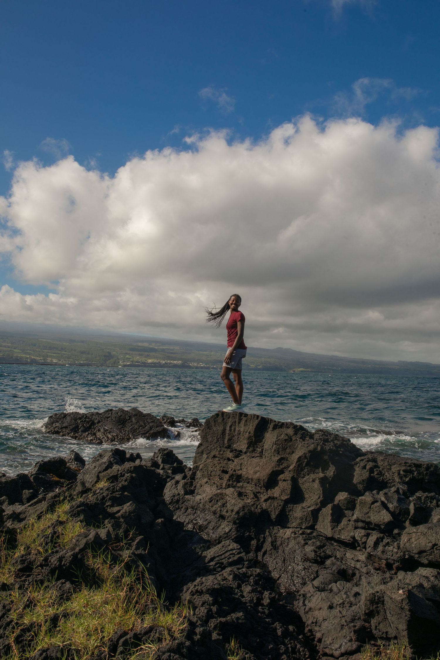 A person poses along rugged volcanic coastline rocks with dramatic cloudy skies and ocean views in Hawaii.