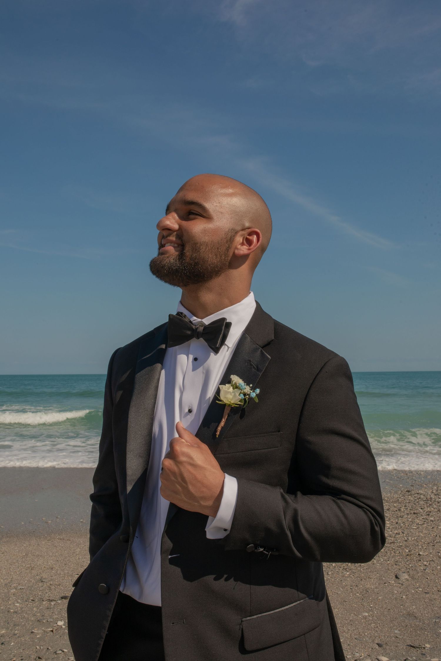 Bald groom in black tuxedo with boutonniere poses confidently on a sunny beach with ocean waves behind him.