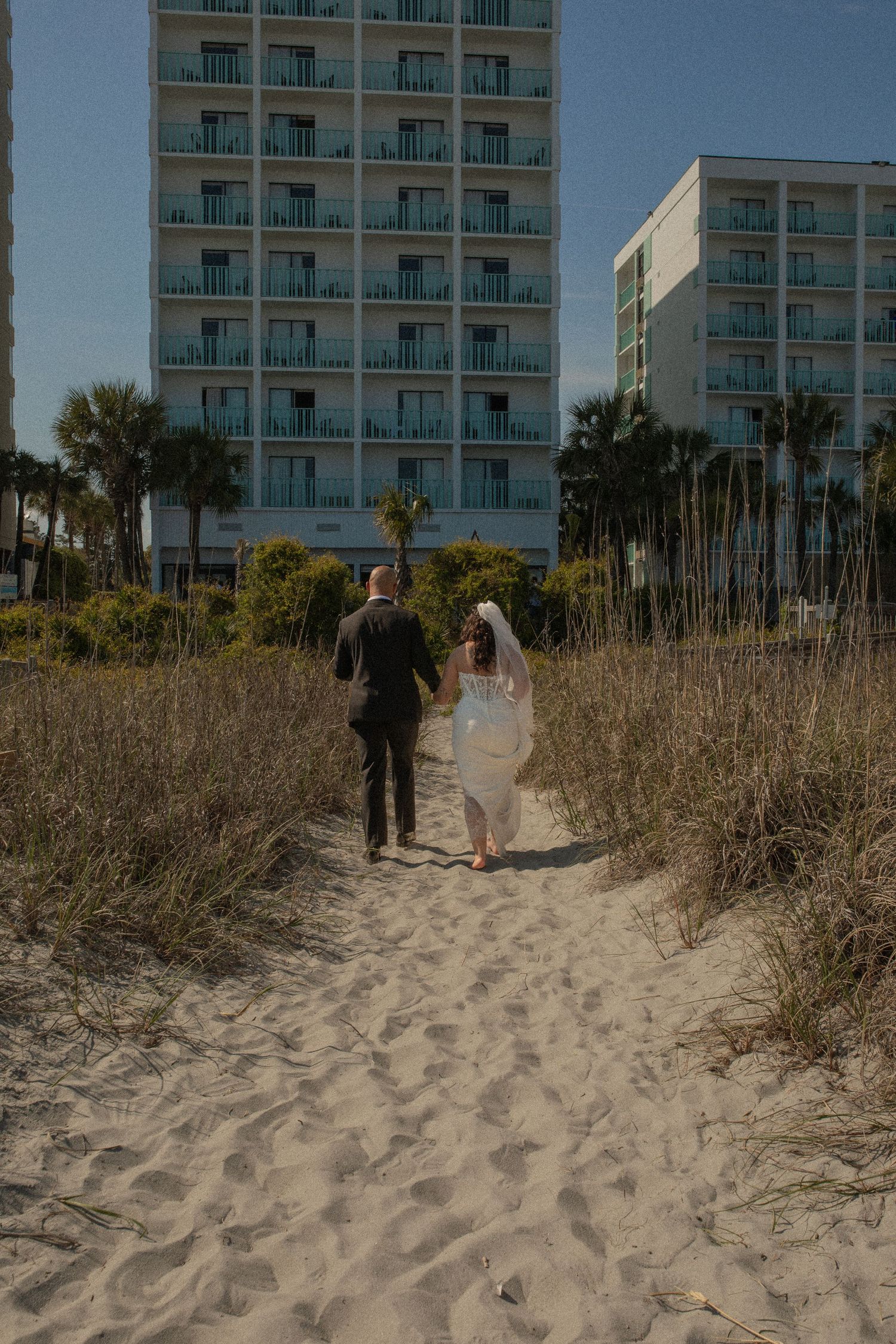 A bride and groom walk hand-in-hand along a sandy beach path toward a coastal hotel surrounded by palm trees.