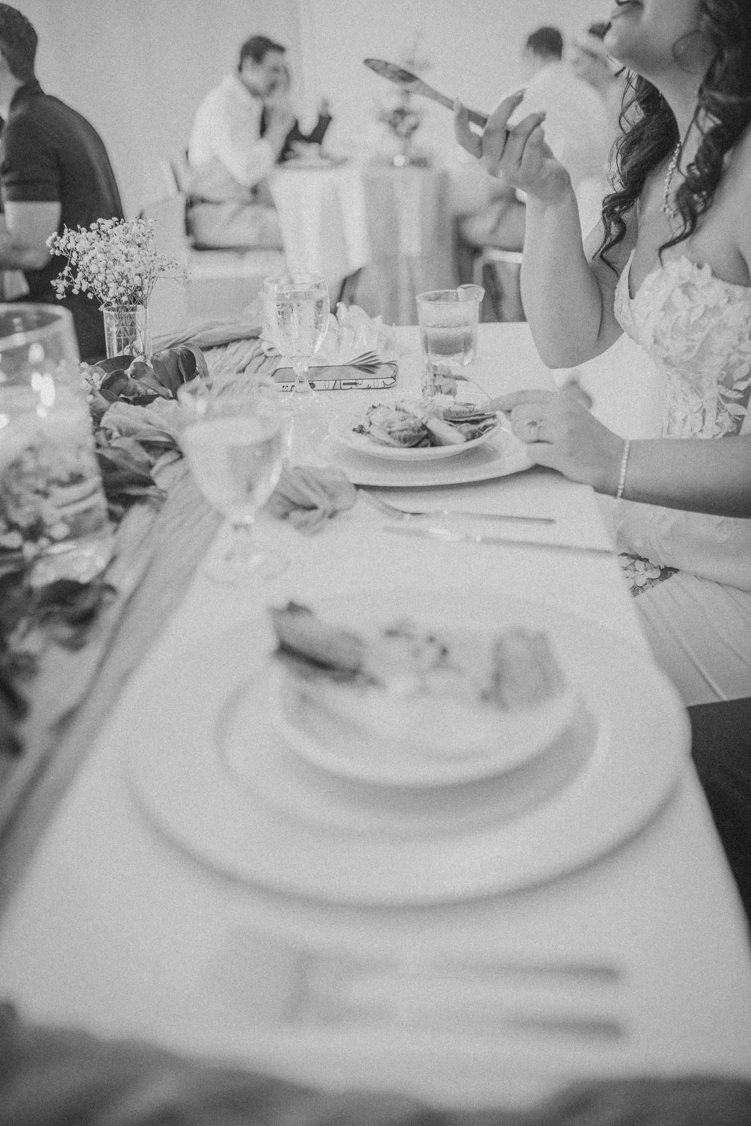 Black and white wedding reception photo showing bride eating at elegantly set table with wine glasses and white plates.