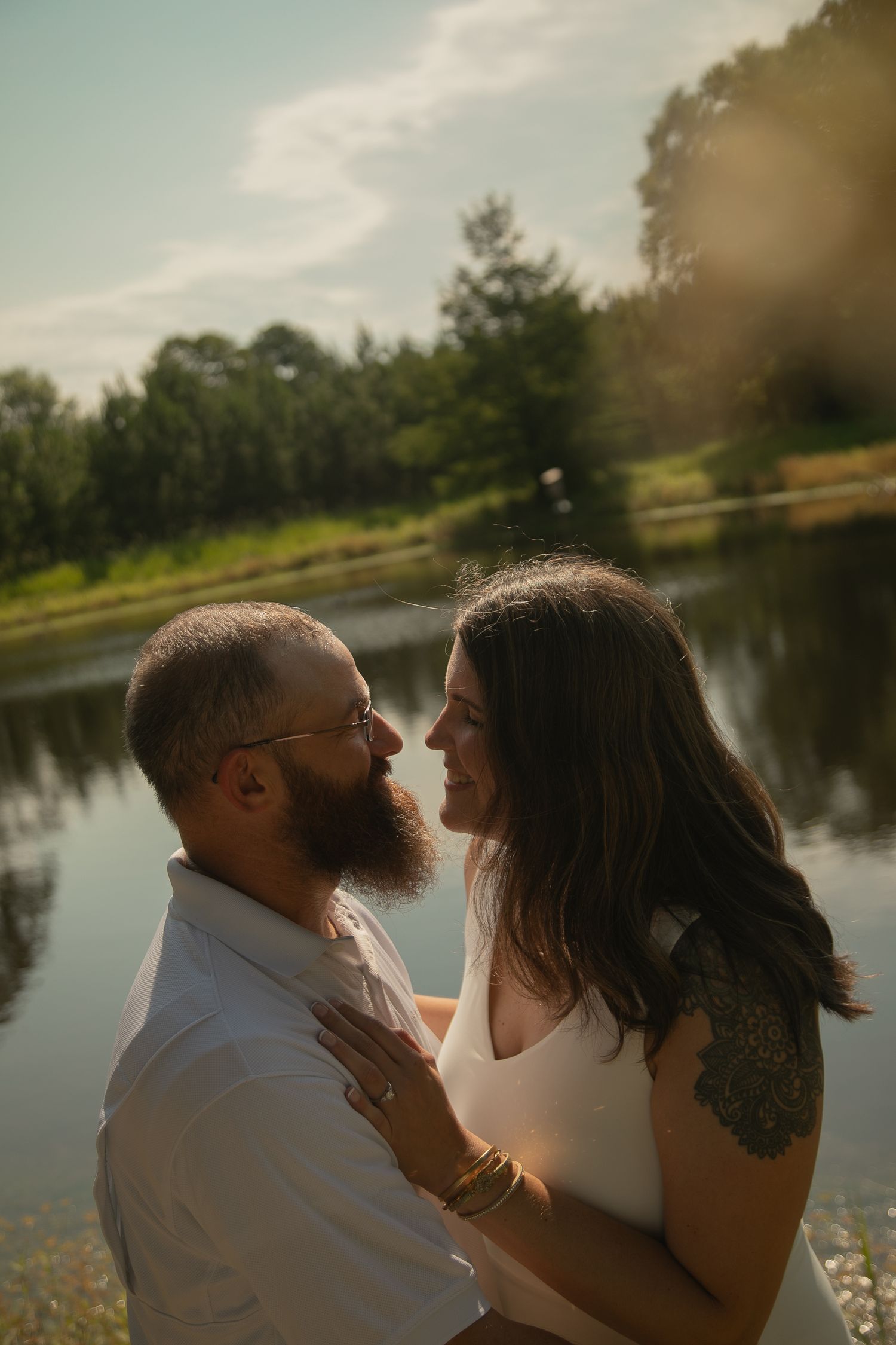 A couple shares a romantic moment by a sunlit pond, the bearded man and tattooed woman gazing at each other lovingly.