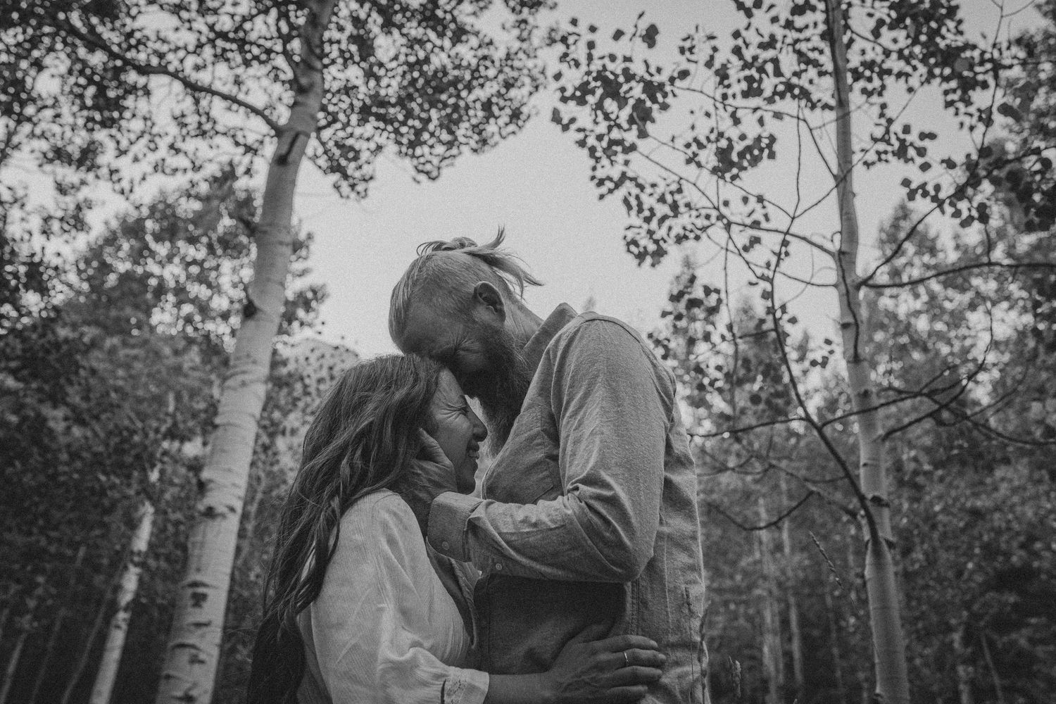 Black and white photo of a romantic couple sharing an intimate moment among tall birch trees in a forest.