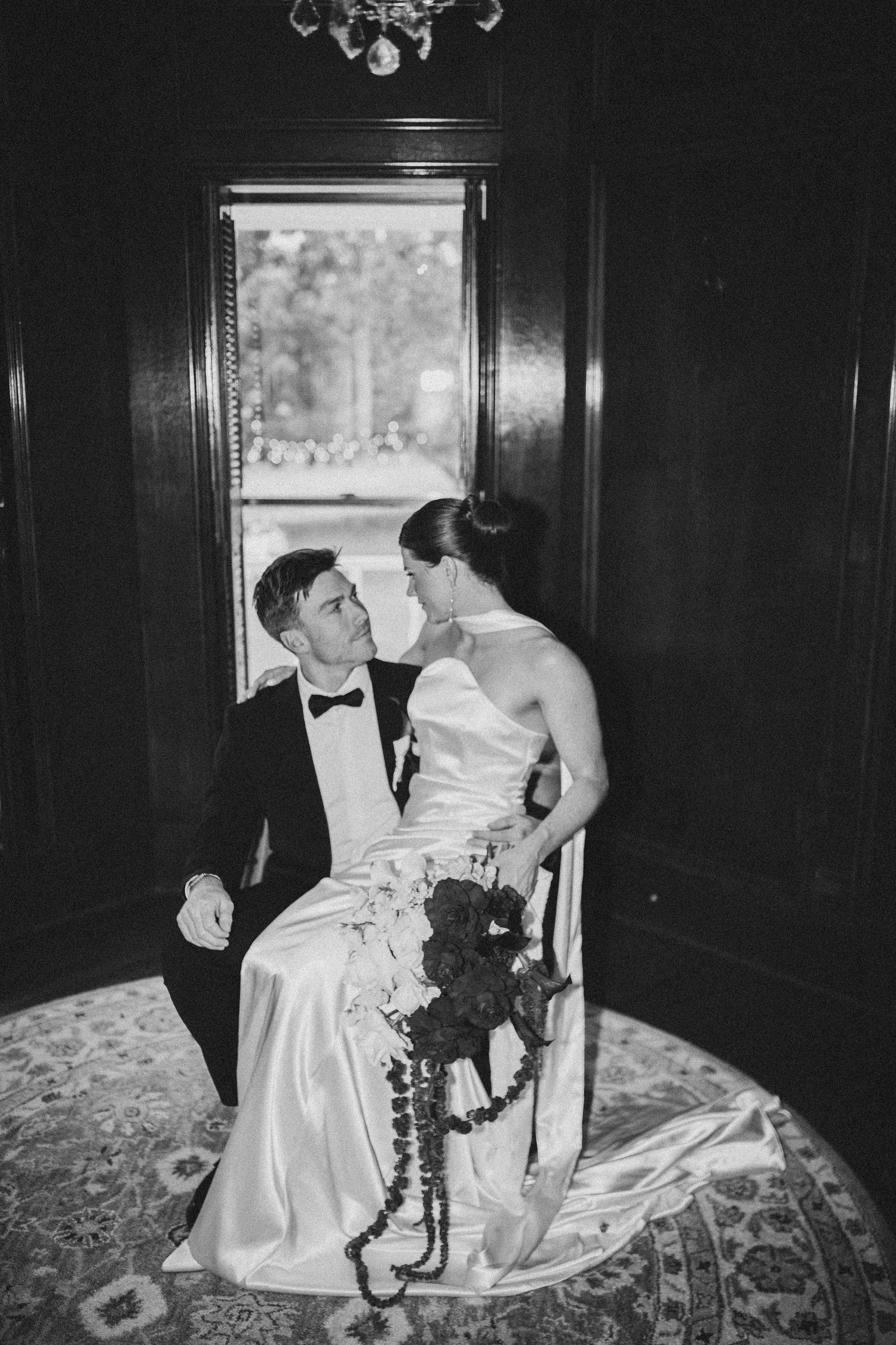 Black and white wedding photo of bride and groom sharing an intimate moment seated by a window in an elegant dark wood room.