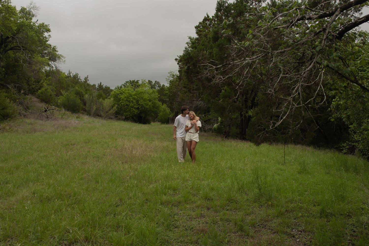 Couple embracing in a lush green meadow surrounded by trees under an overcast sky.