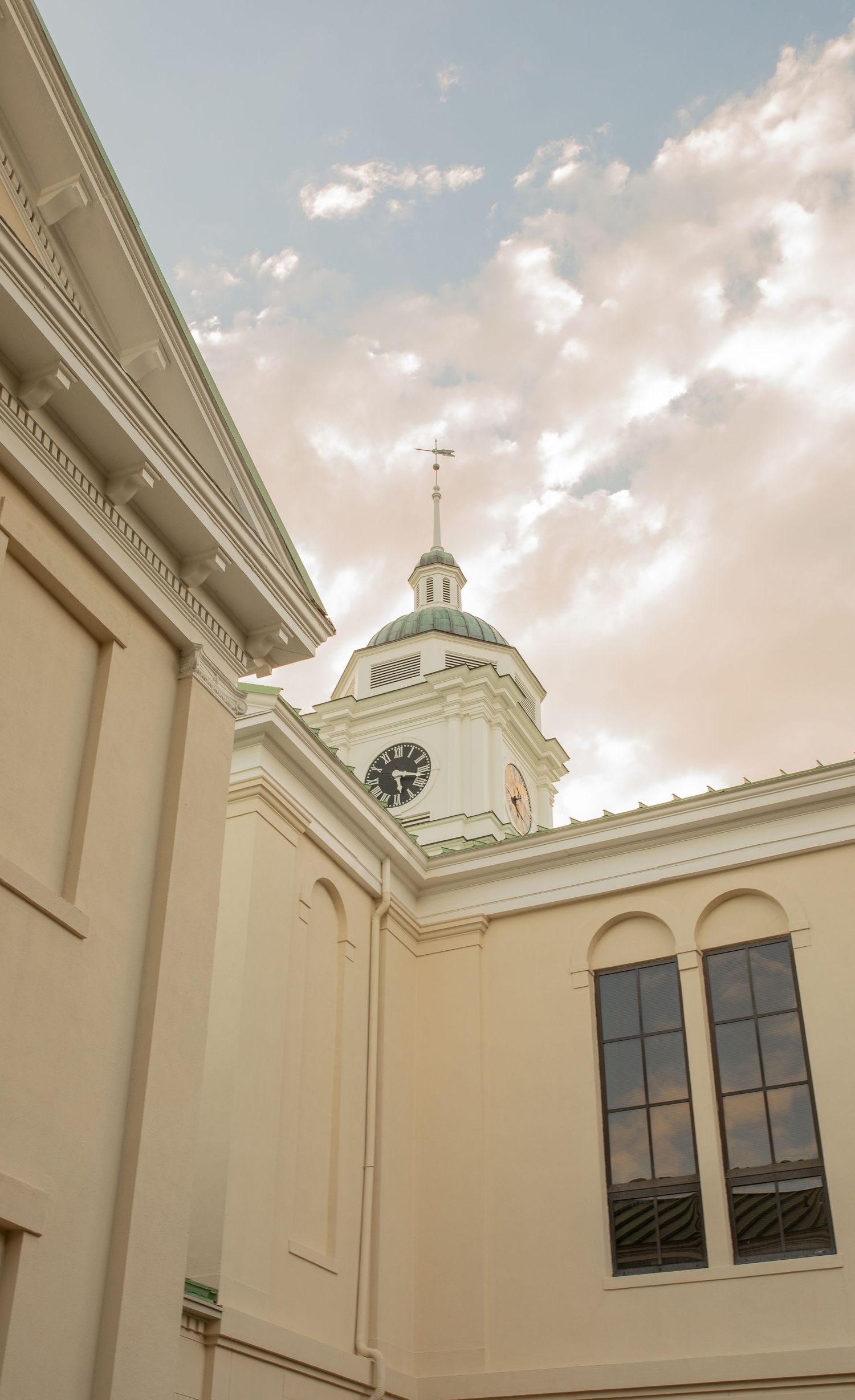 Historic church clock tower with green dome and cross rises above cream-colored neoclassical building under soft cloudy sky.