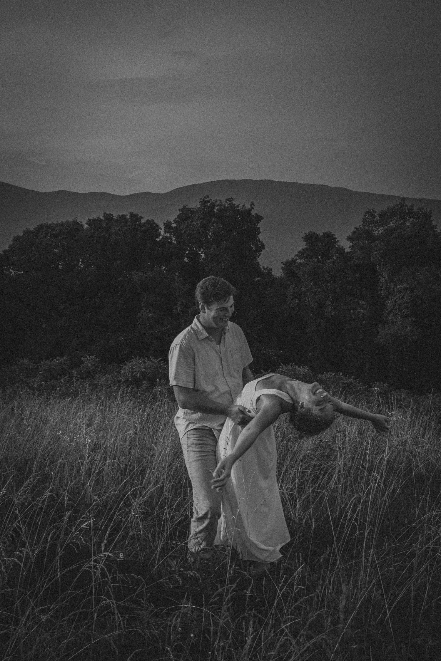Black and white photo of a couple dancing in a field, man dipping woman in white dress, mountains in background.