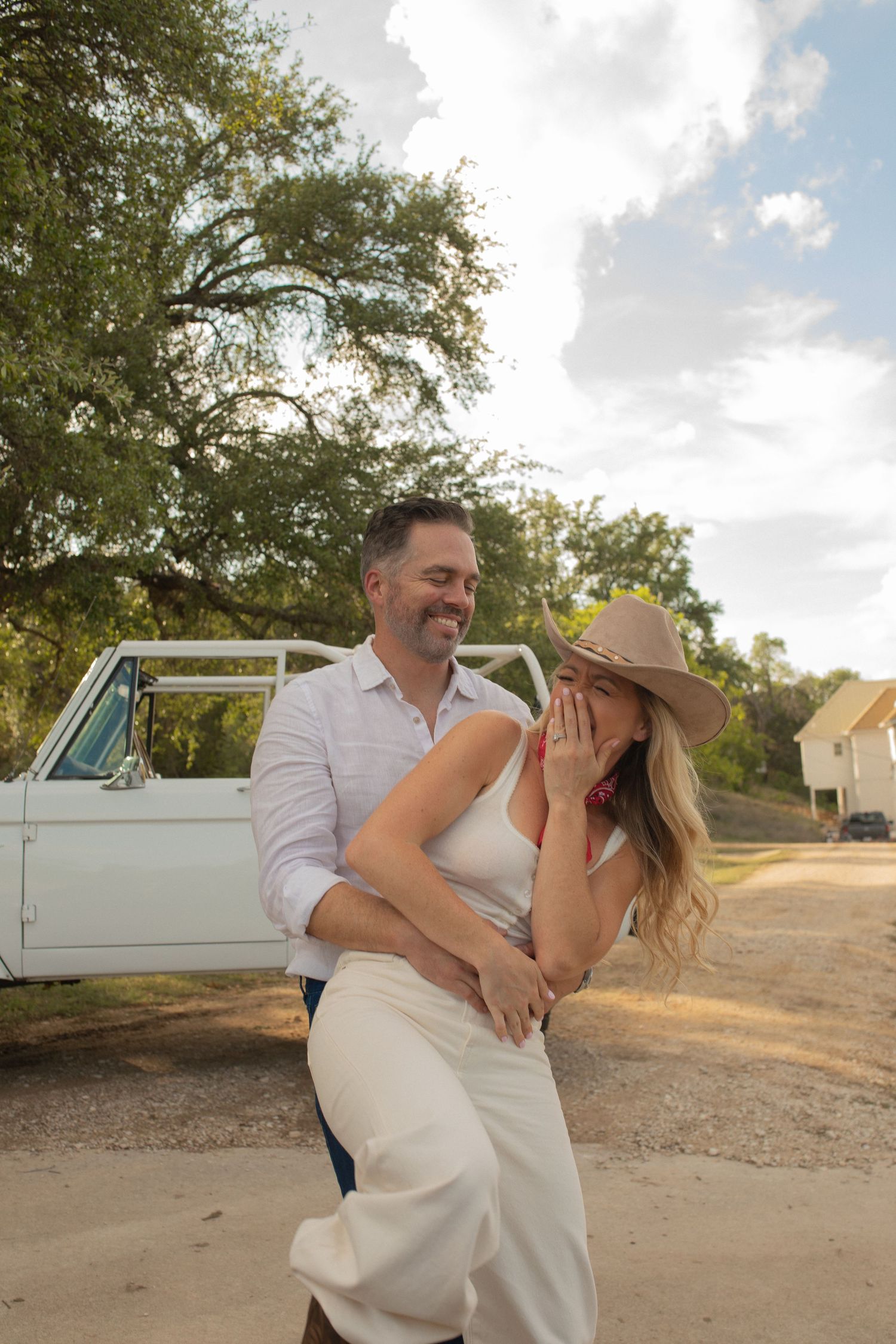 A couple embraces outdoors near a vintage white truck, woman in cowboy hat and white outfit, man in white shirt smiling.