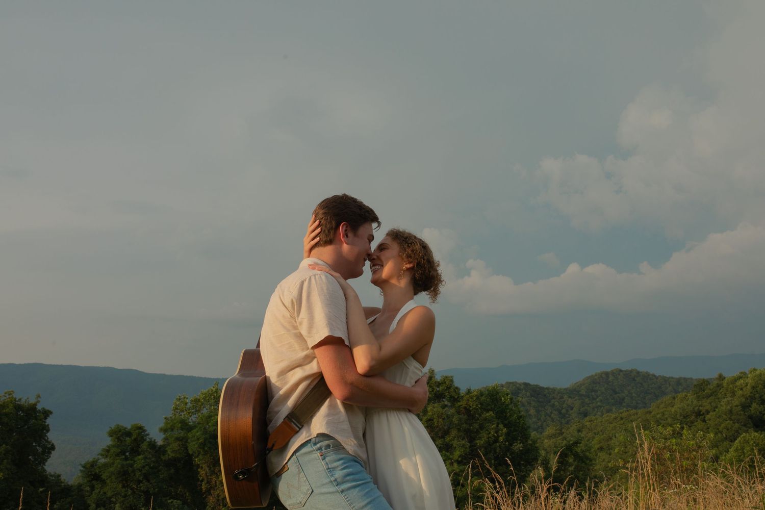 Romantic couple sharing a tender moment outdoors with a guitar, rolling hills and dramatic sky in the background.