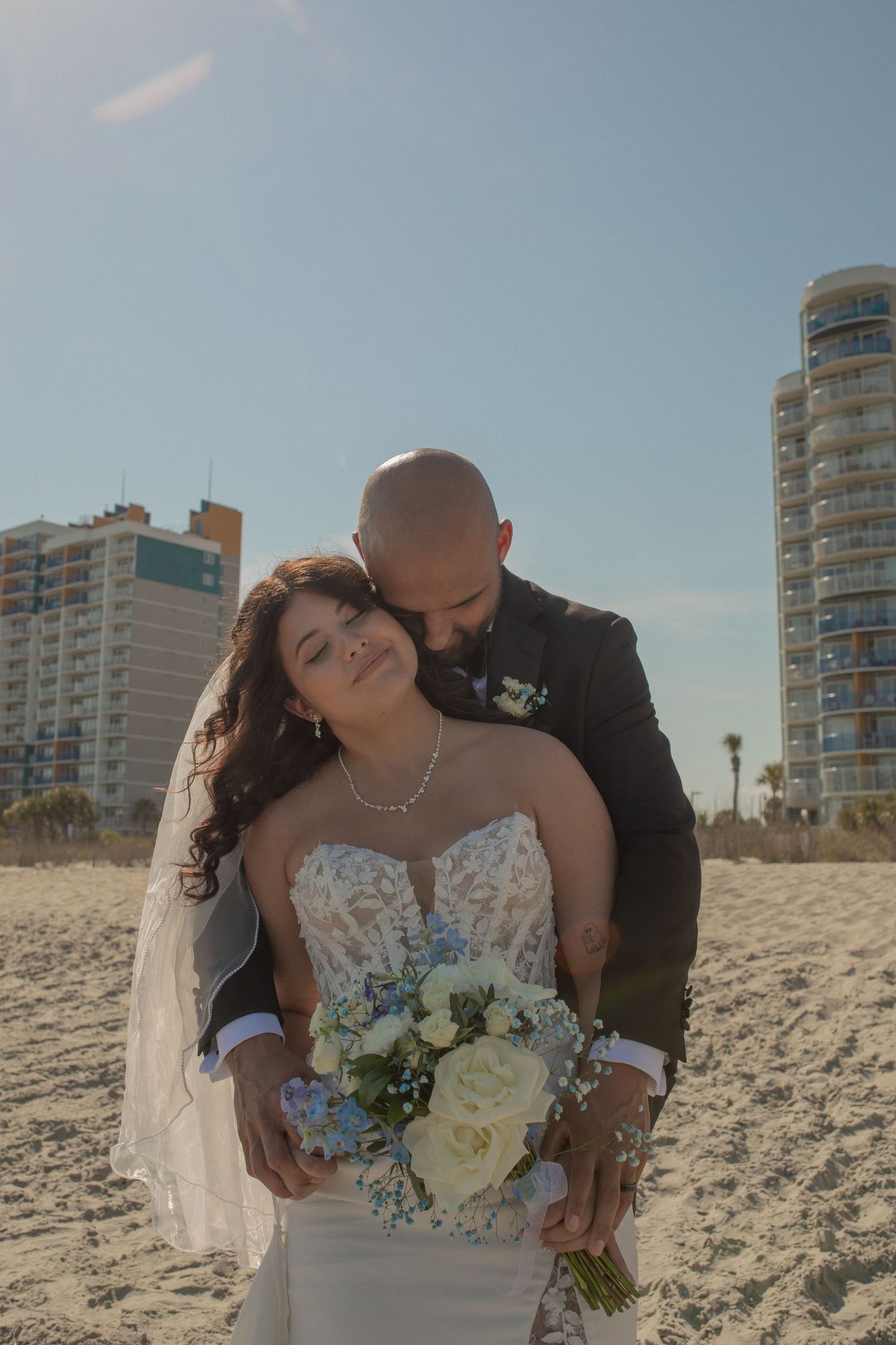 Bride and groom embrace on a sunny beach, bride holding blue and white bouquet, tall buildings in background.
