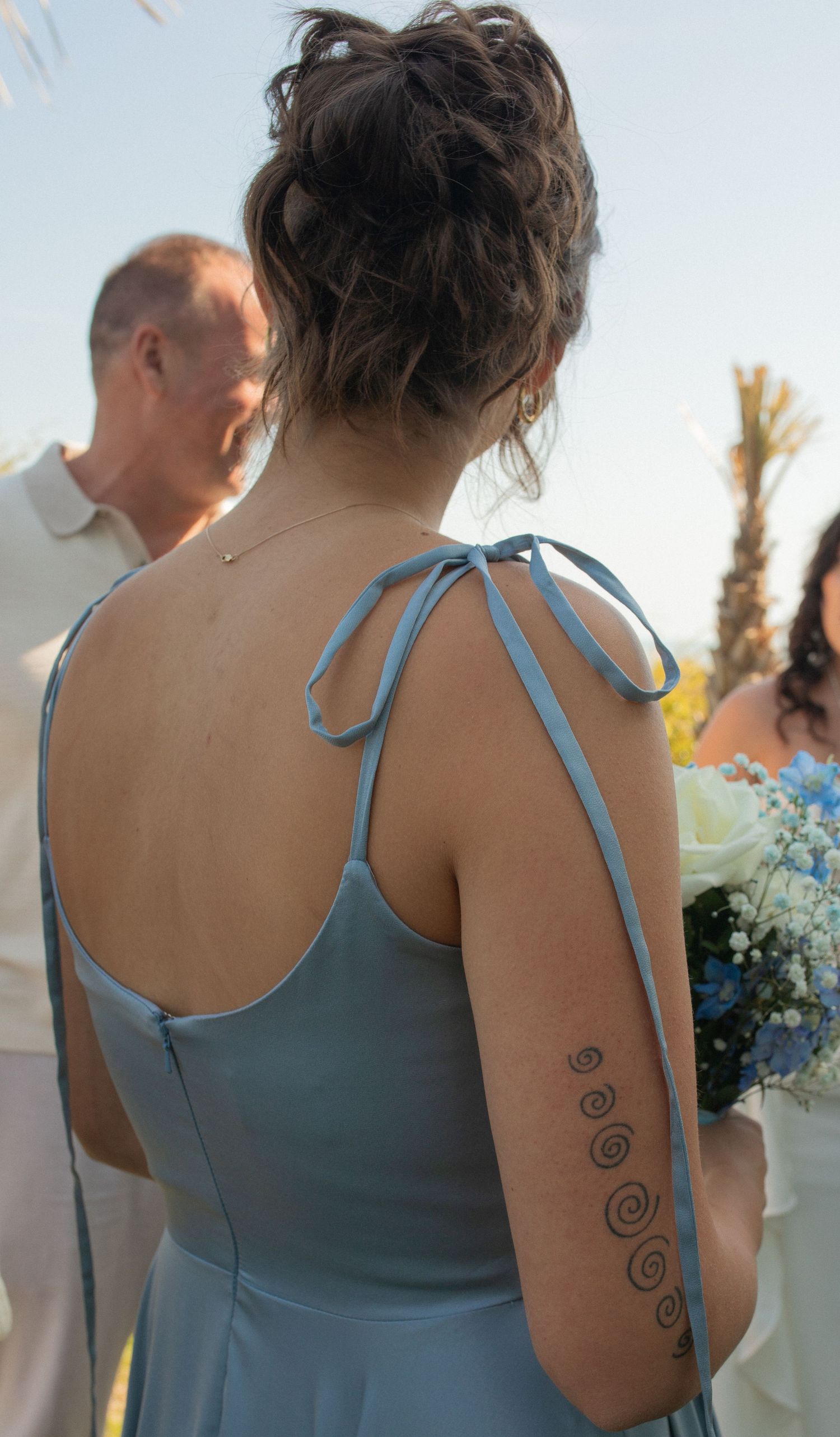 Bridesmaid in light blue dress with spiral tattoo on arm holds bouquet at outdoor wedding ceremony.