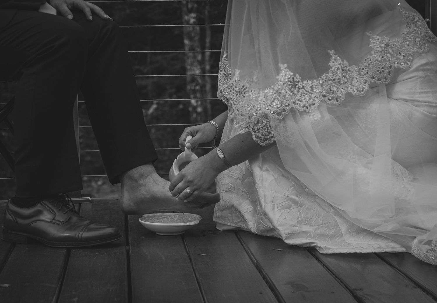 Black and white photo of bride and groom during wedding foot washing ceremony on wooden deck.