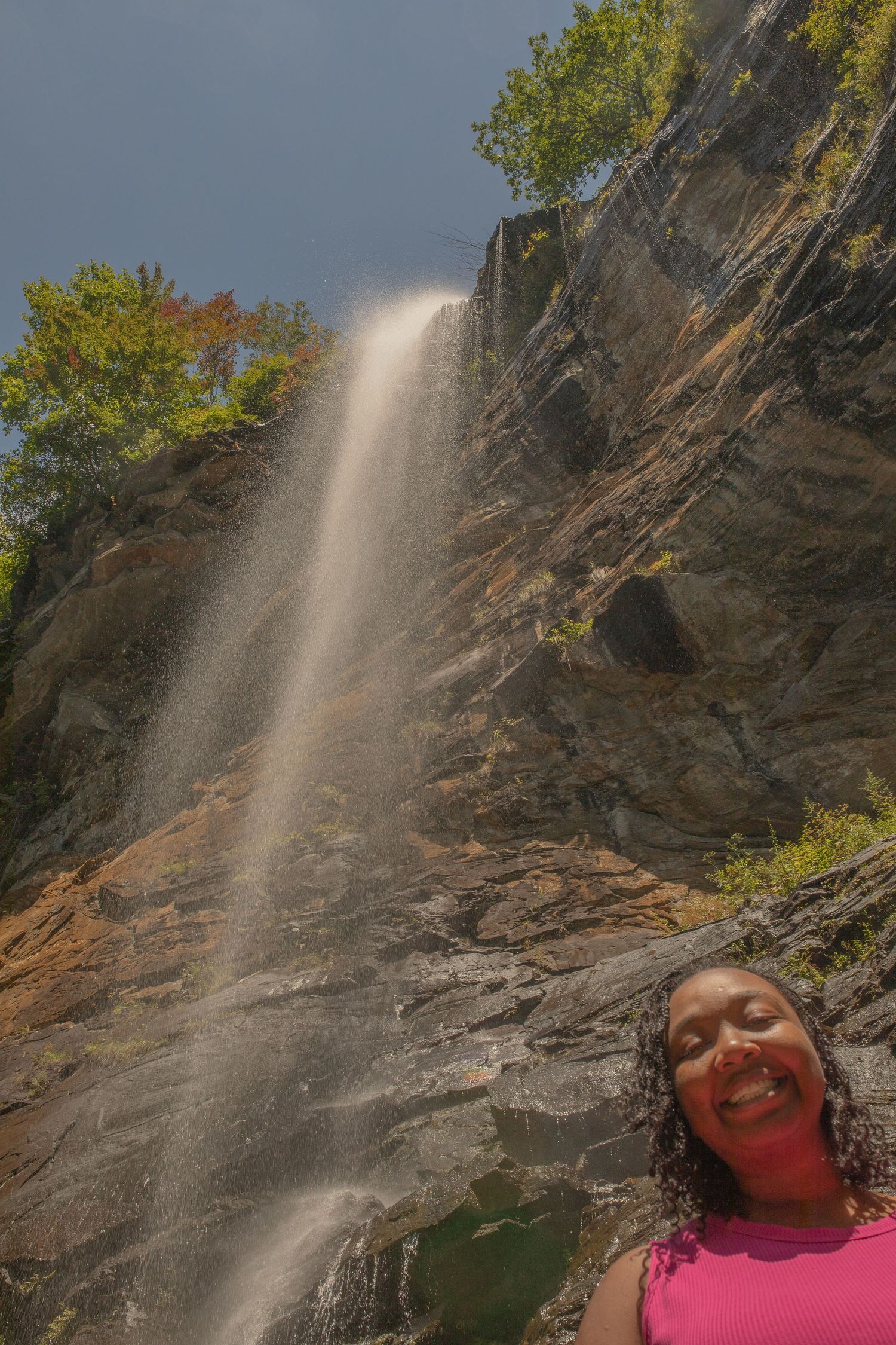 Woman smiling in front of a tall waterfall cascading down rocky cliffs surrounded by lush green trees.