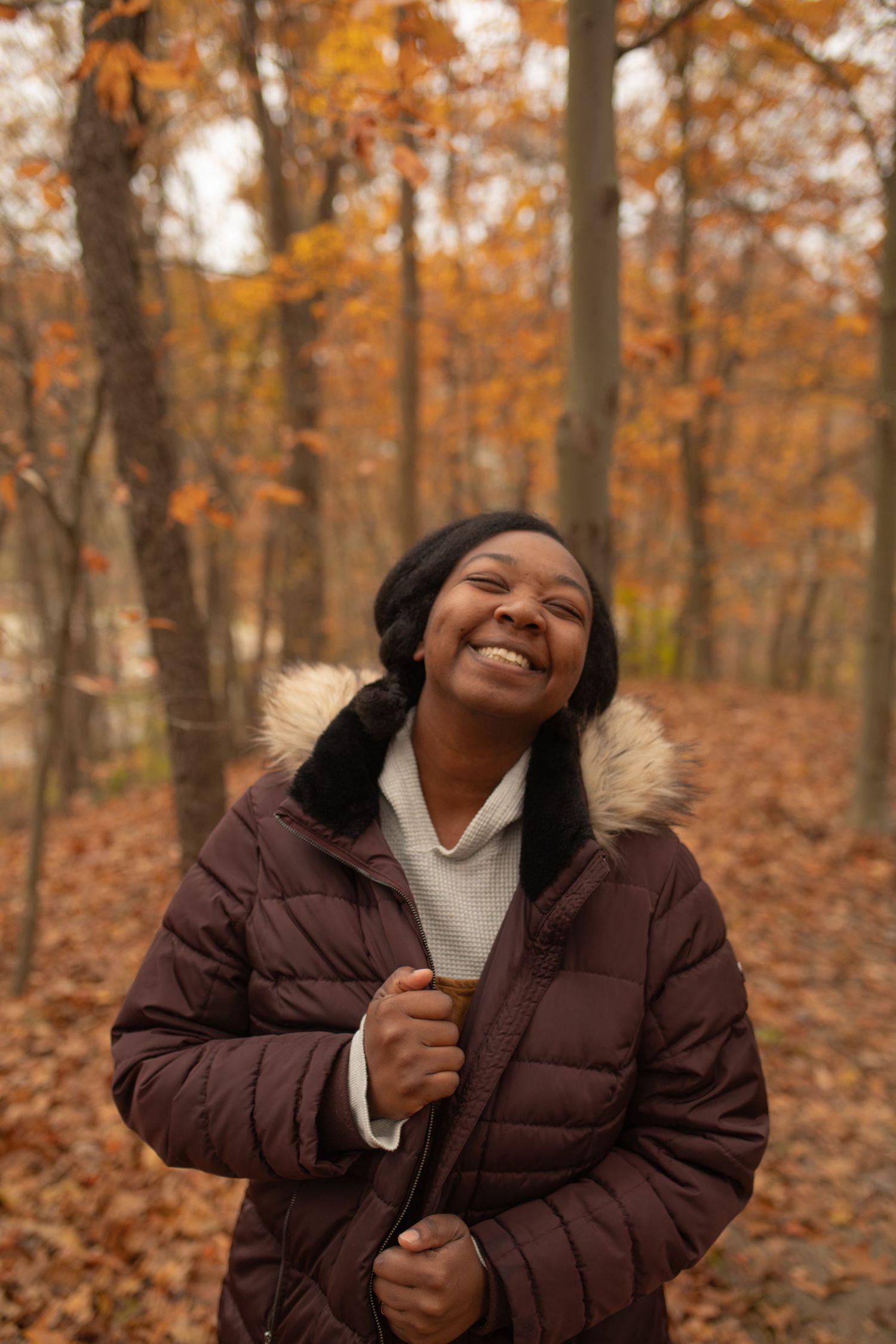 A smiling young Black woman in a brown fur-hooded puffer jacket stands in an autumn forest with orange foliage.