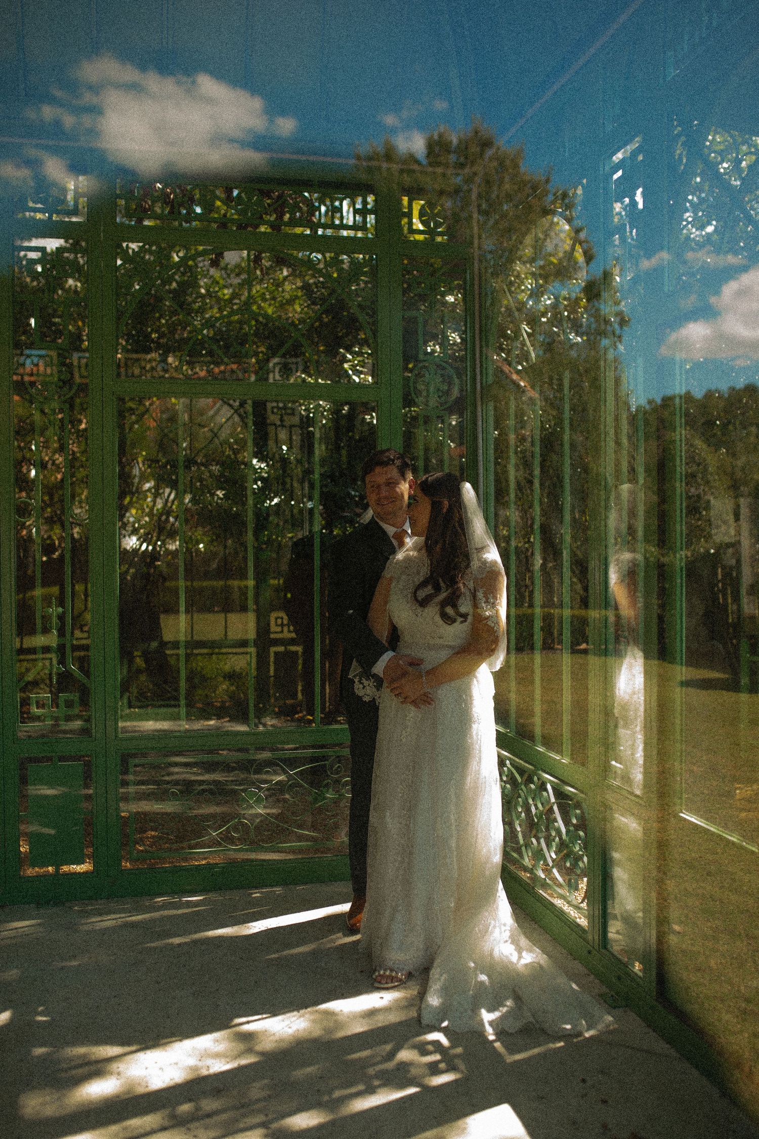 A couple shares a romantic moment in a sunlit garden gazebo with ornate green metalwork and natural light filtering through.