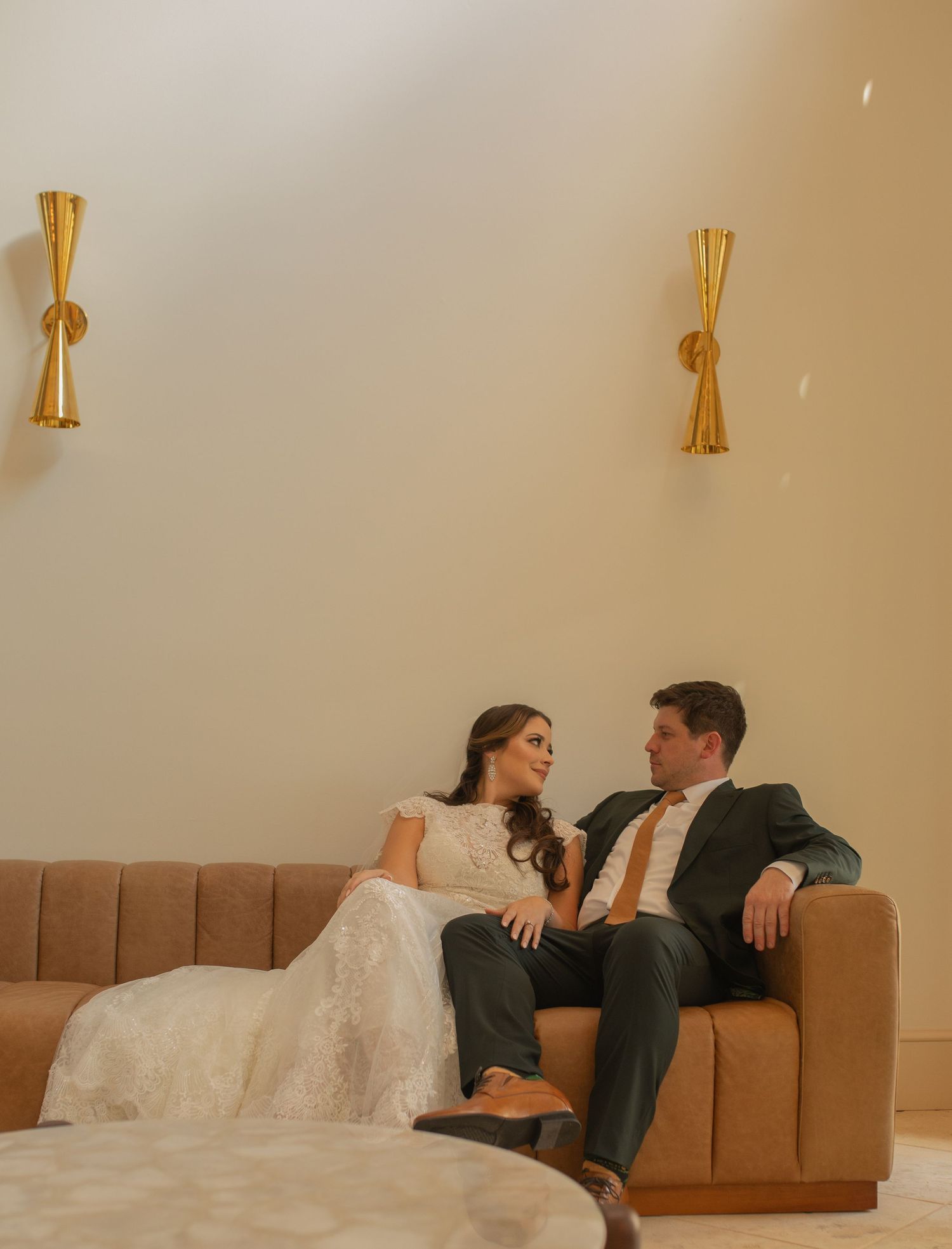 A couple in wedding attire relaxes on a tan leather couch with gold wall sconces in the background.