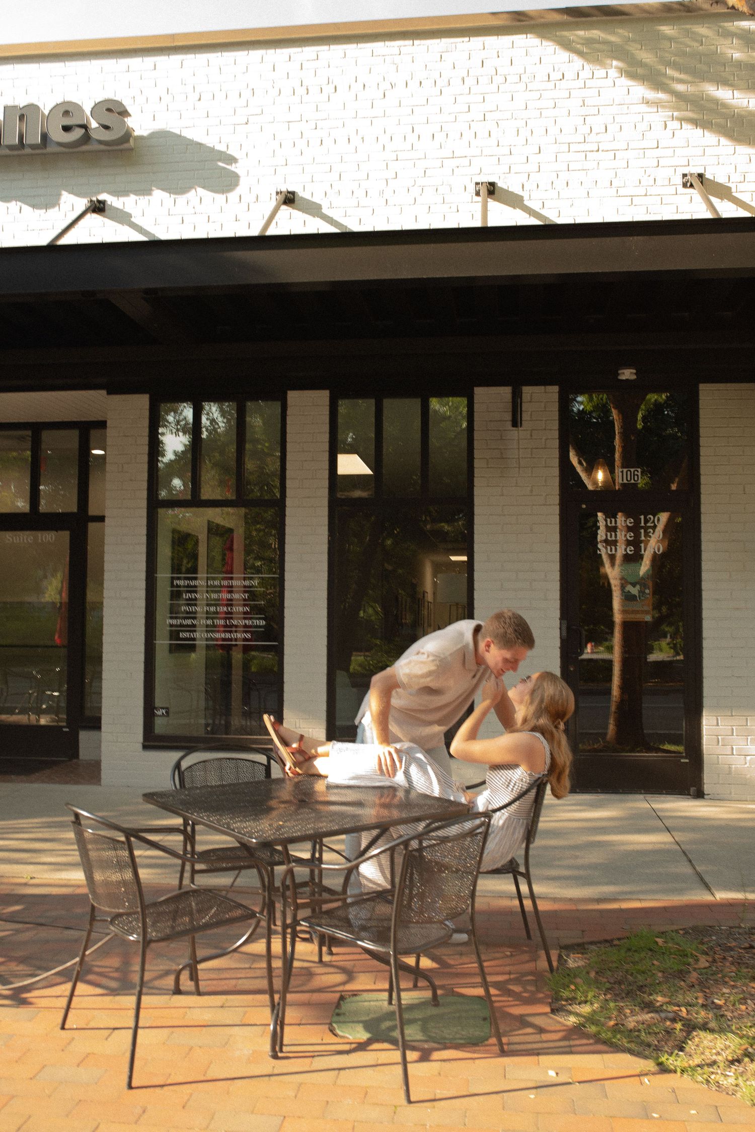 Couple sharing an intimate moment at an outdoor cafe table on a sunny afternoon near a modern storefront.
