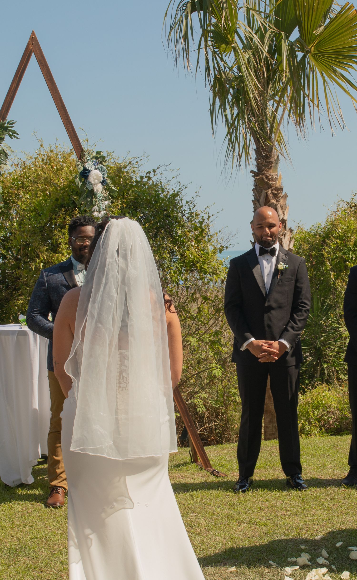 Outdoor wedding ceremony with bride in white veil approaching groom in black suit near triangular arch with palm trees.
