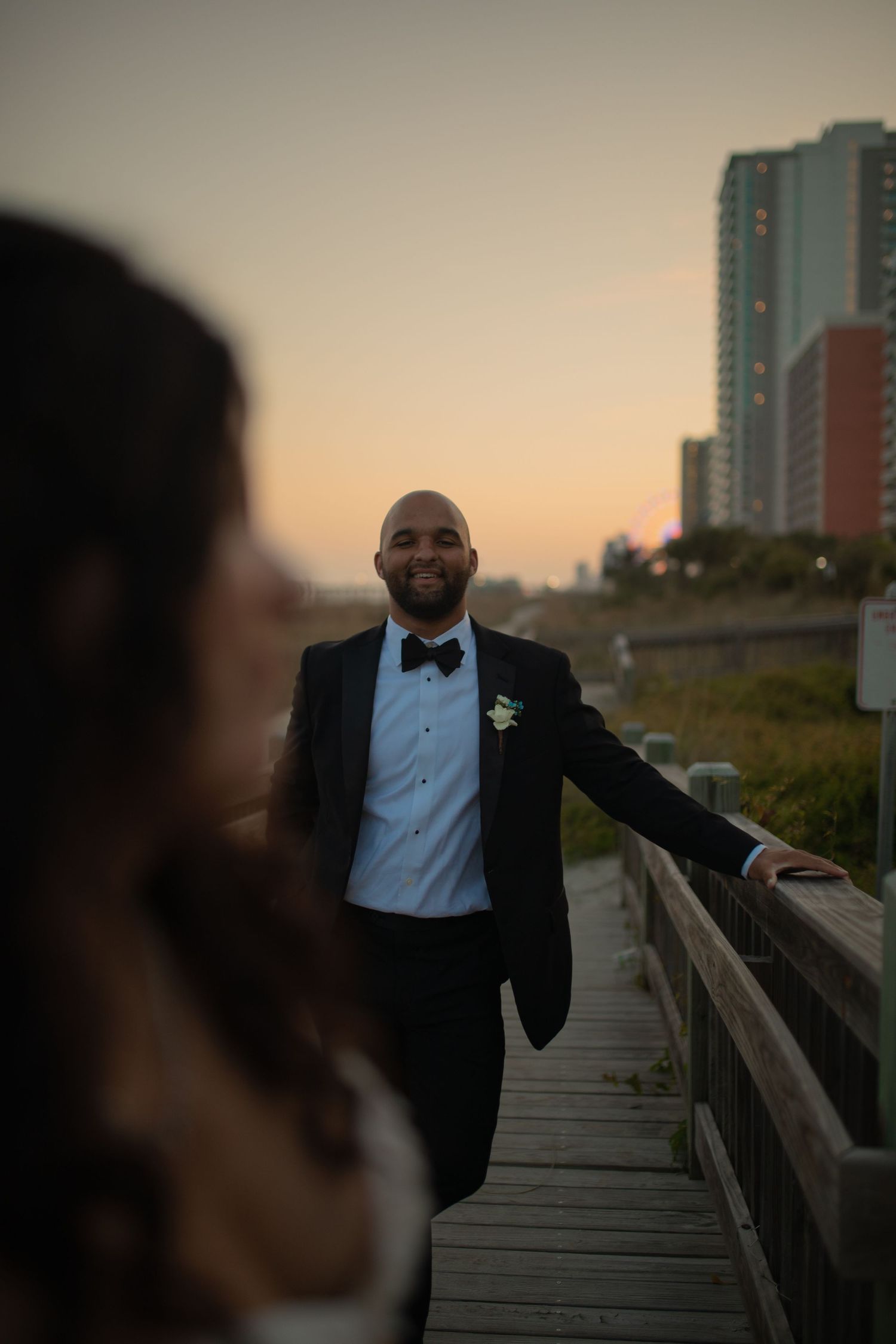Groom in black tuxedo with boutonniere smiles on boardwalk at sunset, bride blurred in foreground.