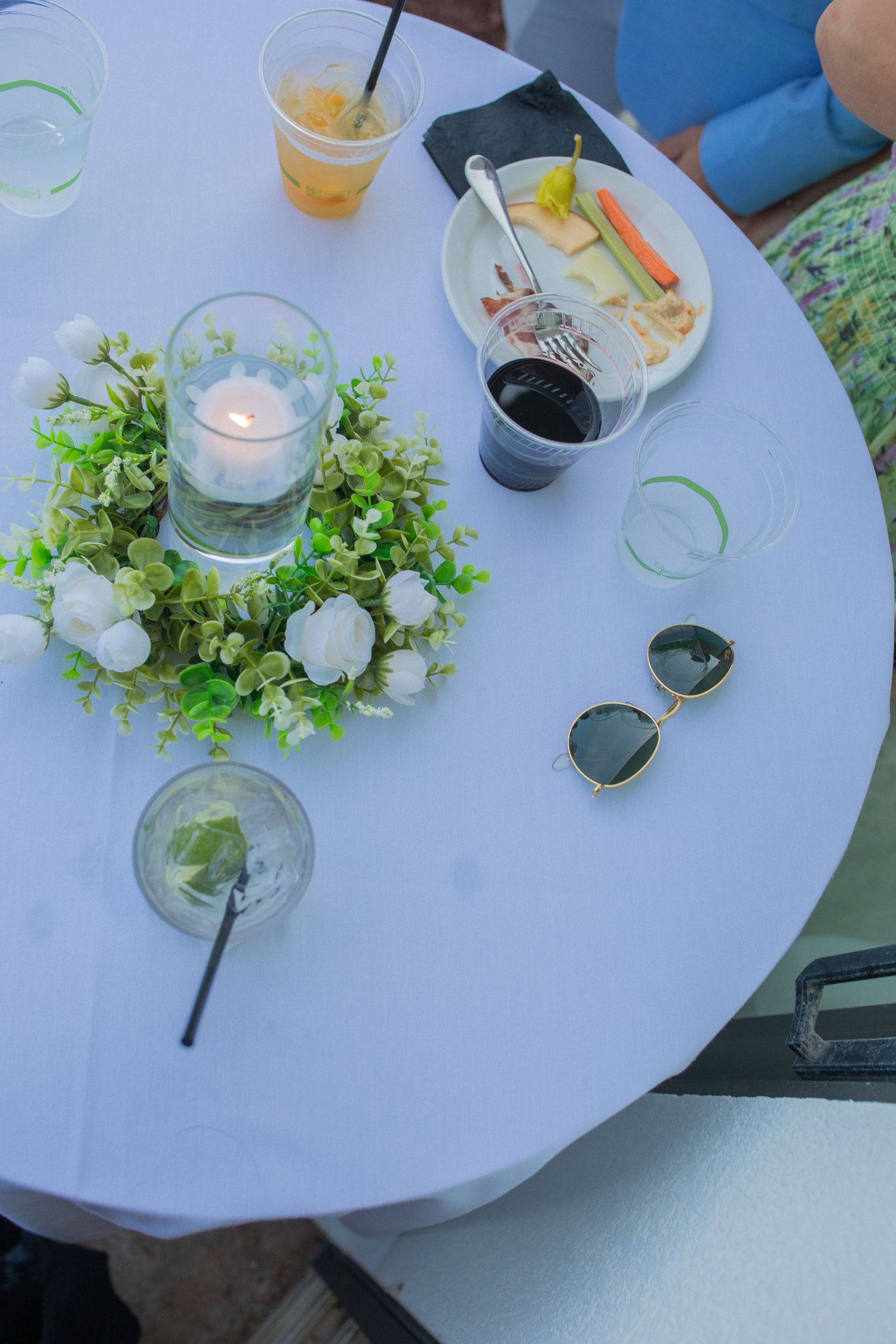 White round table with floral candle centerpiece, sunglasses, drinks, and appetizer plate at an outdoor event.