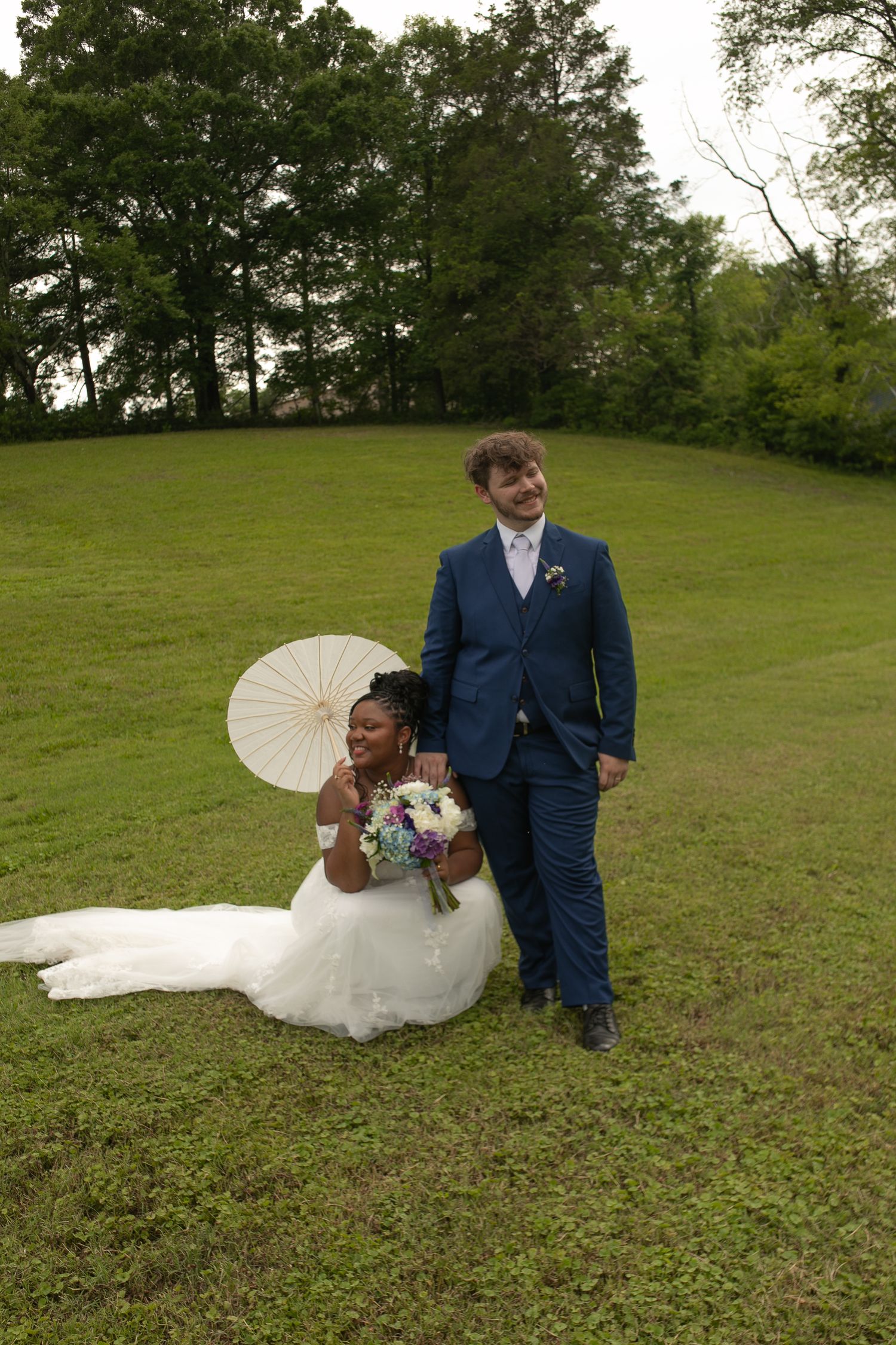 Bride in white gown with parasol sits on grass beside groom in blue suit holding bouquet at outdoor wedding.