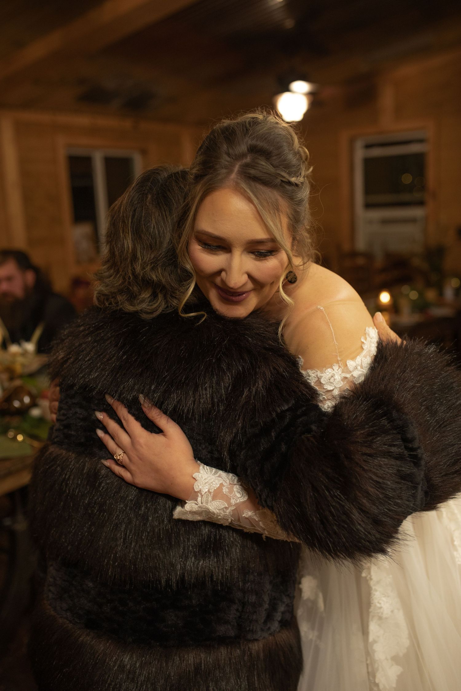 Bride in white lace gown hugging a guest in dark fur coat at a warm, rustic wedding reception venue.
