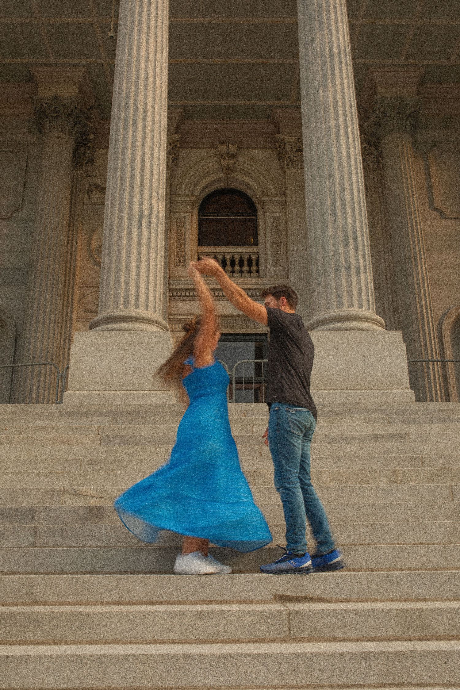 Couple dancing on steps of a grand building with columns, woman in flowing blue dress being twirled by man in jeans.