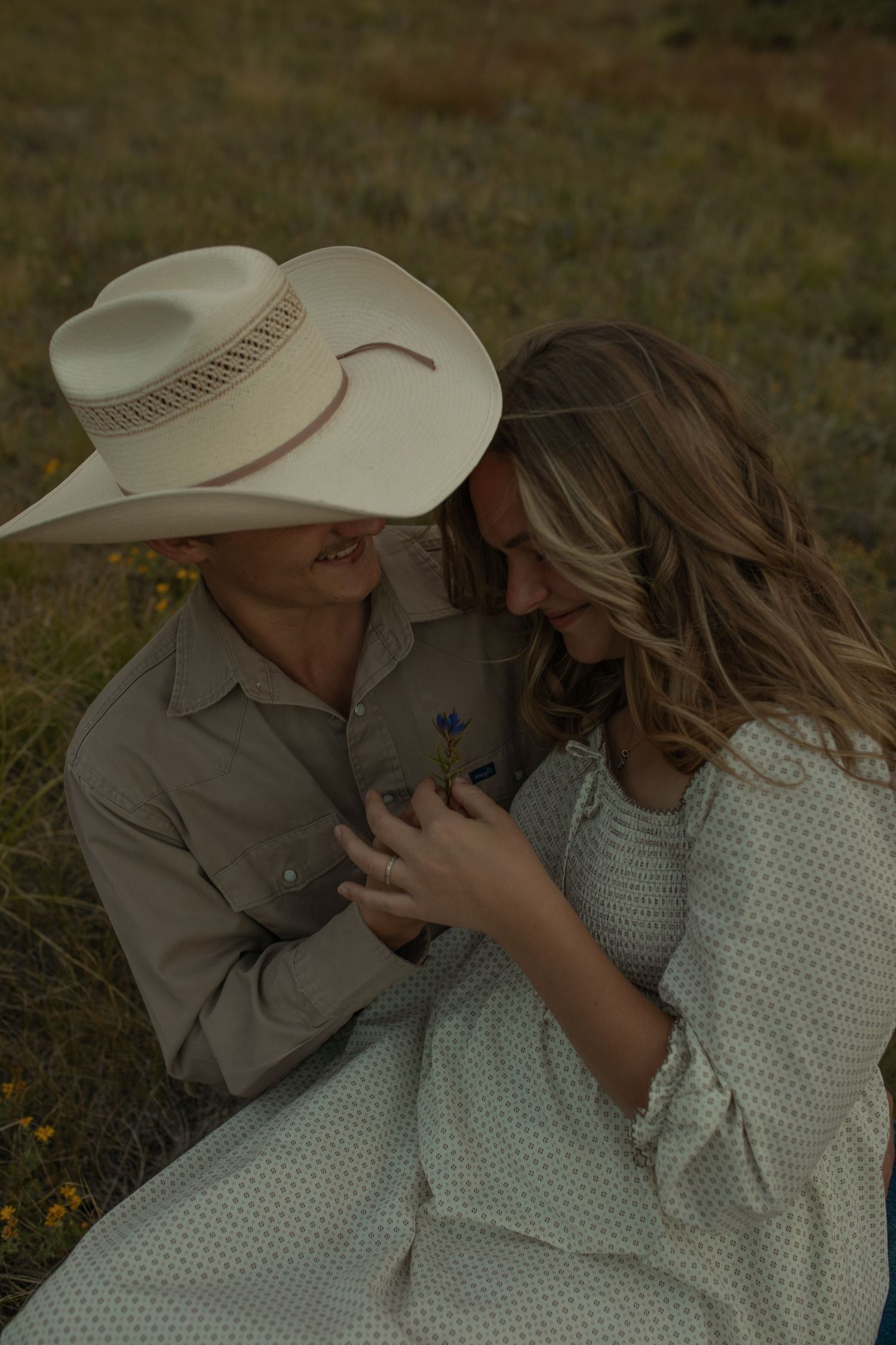 A romantic couple in western attire share tender moments while sitting together in a grassy field.