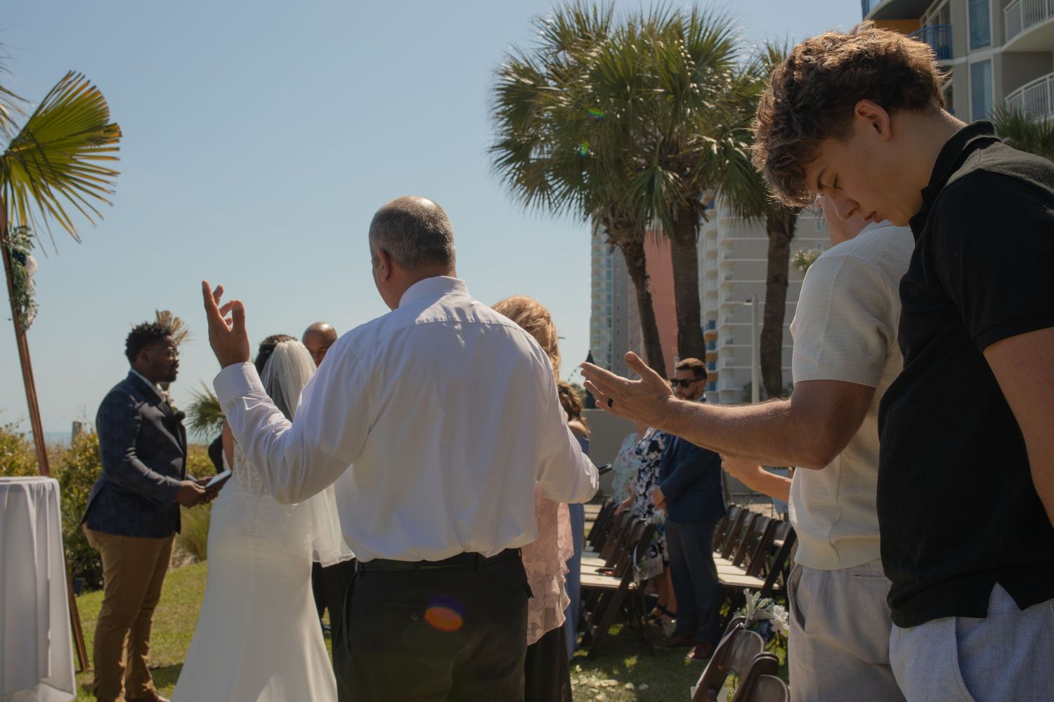Outdoor wedding ceremony on sunny day with guests gathering near palm trees and a tall building in background.