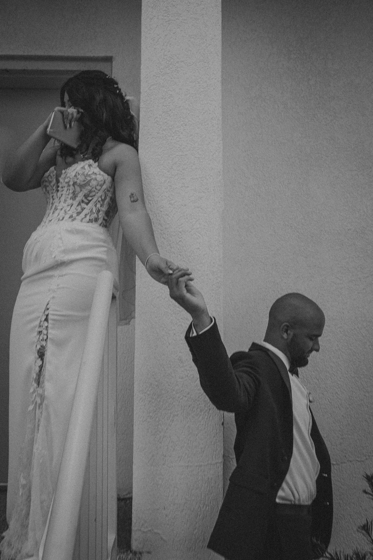 Black and white wedding photo of bride in lace gown holding groom's hand as he kneels beside a wall.