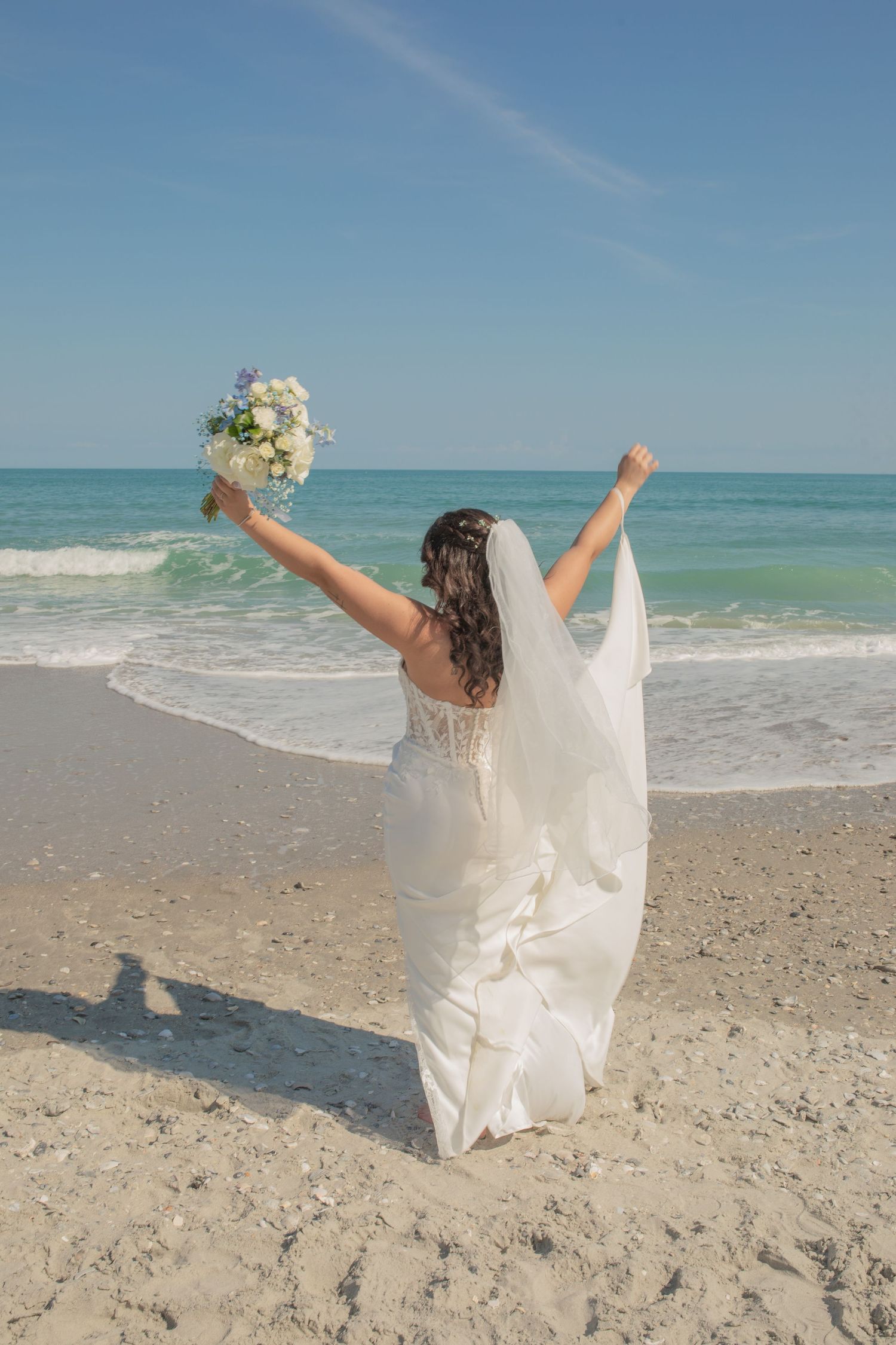 Bride in white gown and veil celebrating on a sandy beach, arms raised holding a bouquet, with turquoise ocean waves behind her.